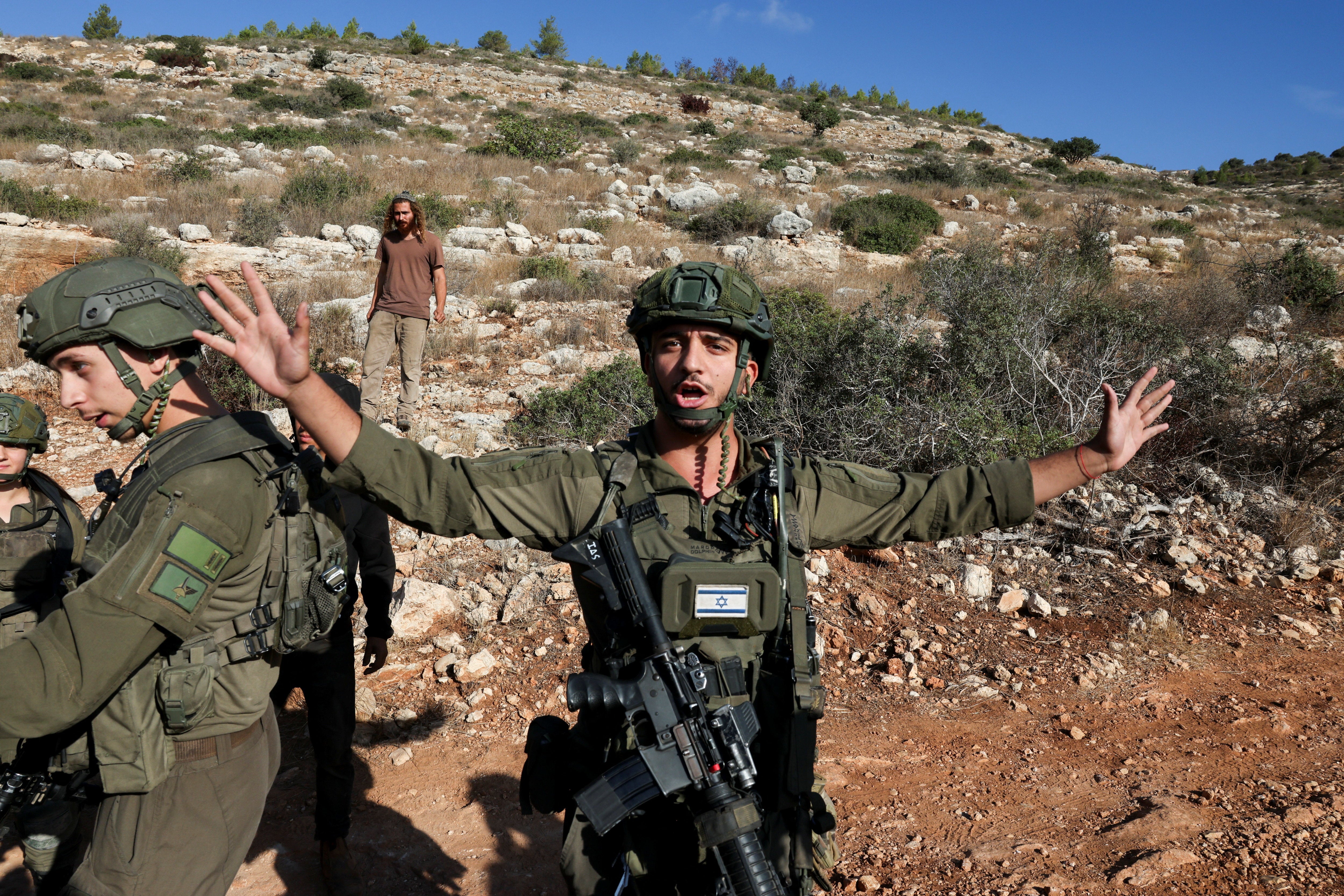 A member of the Israeli forces gestures as an Israeli settler walks in the background during the olive harvest at Palestinian-owned olive groves in Deir Abu Mashal, near Ramallah, in the Israeli-occupied West Bank, October 19, 2026