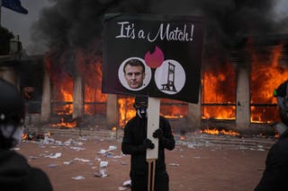 A protester holds a placard with a photo of French president Emmanuel Macron and a guillotine in front of a burning tourism office during a demonstration against rising living costs and in solidarity with students' grievances in Lyon