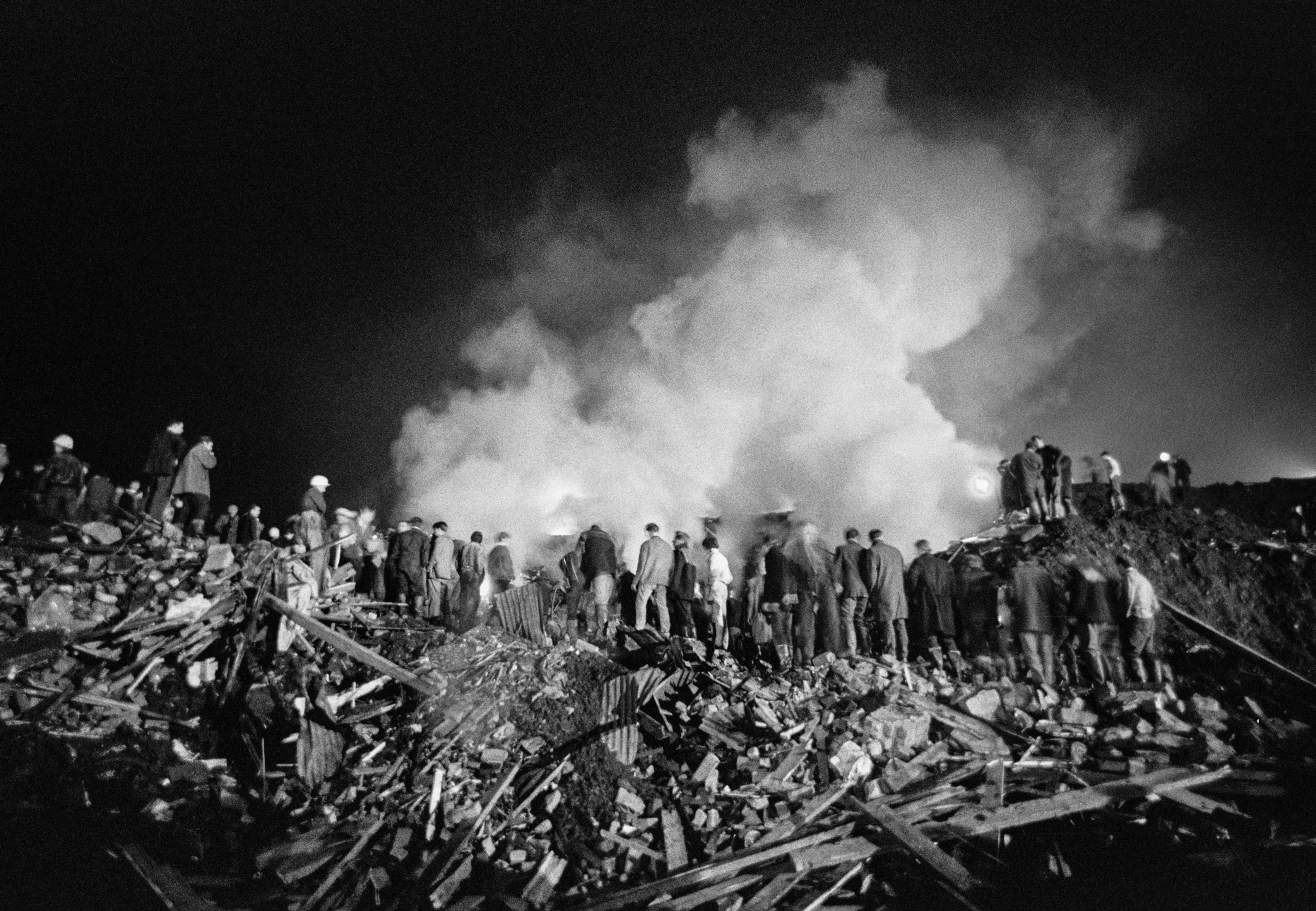 Rescue workers stand on a mound of rubble amid smoke at night in the aftermath of the Aberfan Disaster,
