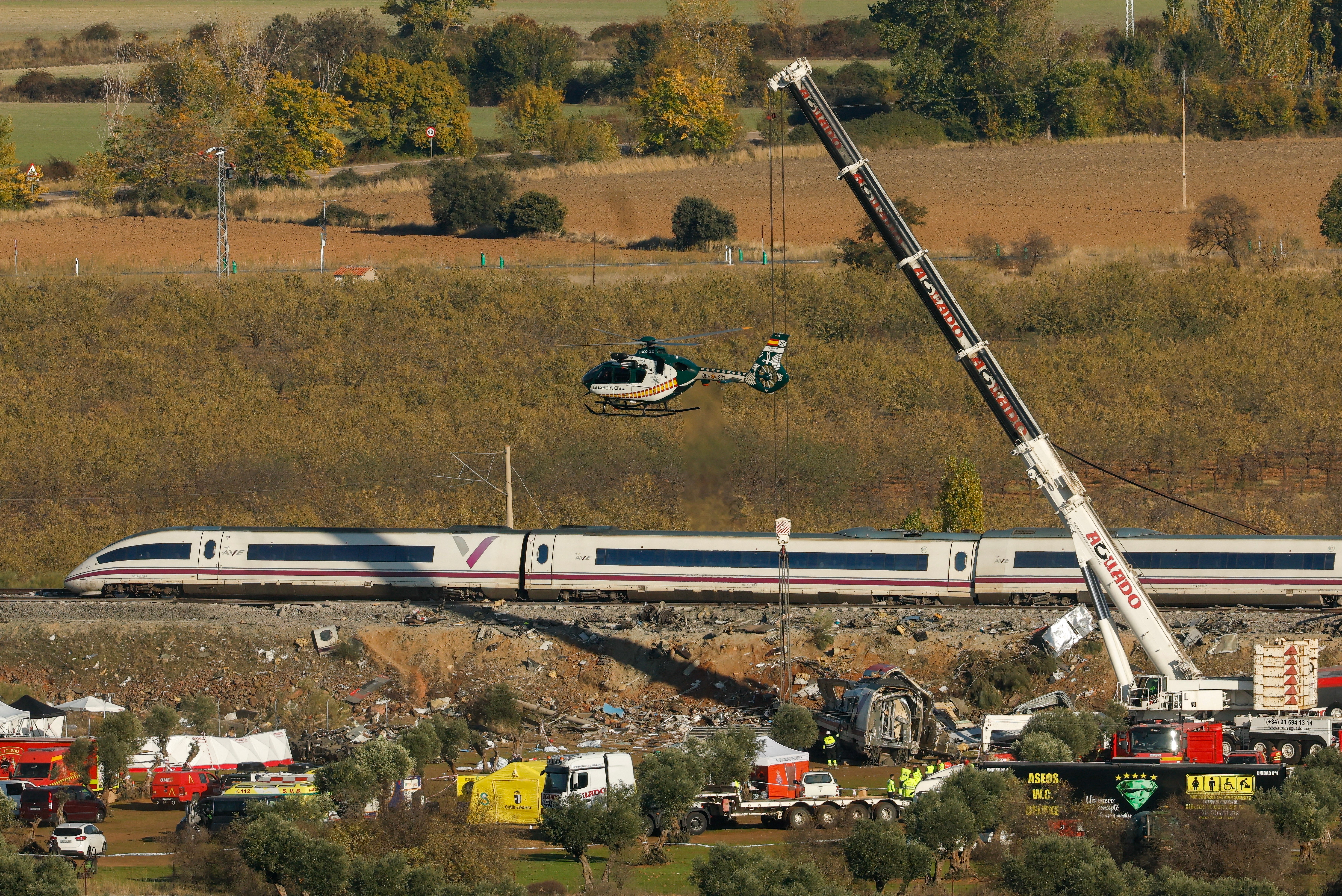 A Civil Guard helicopter hovers as emergency crews use heavy cranes to remove damaged carriages of a crash between two high-speed trains