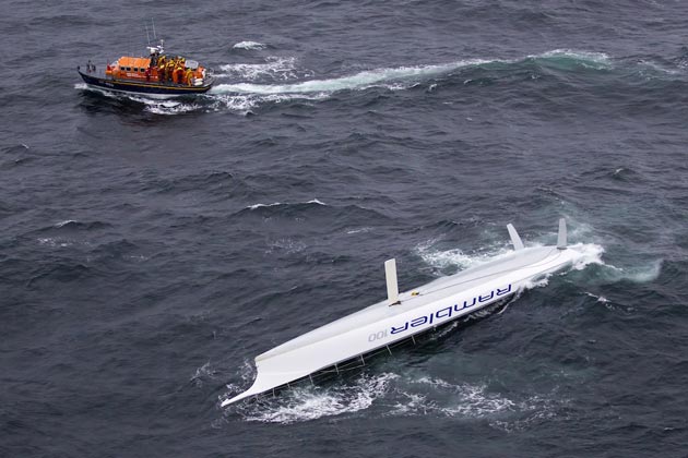 The upturned 100-foot Rambler, with the Baltimore lifeboat in the background, after capsizing just south of the Fastnet rock and lighthouse in the Rolex Fastnet Race