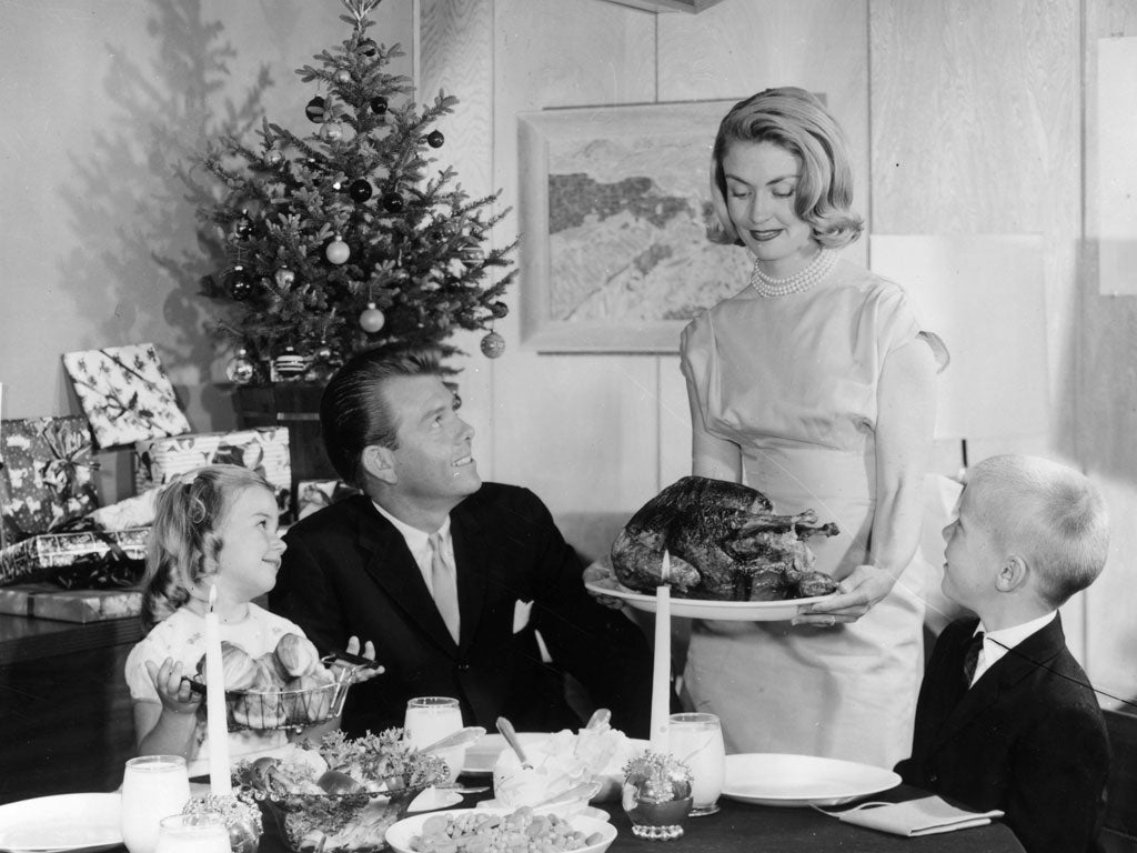 1965: An American family admire a turkey their mother is bringing to the table. A small Christmas tree with presents is in the background.