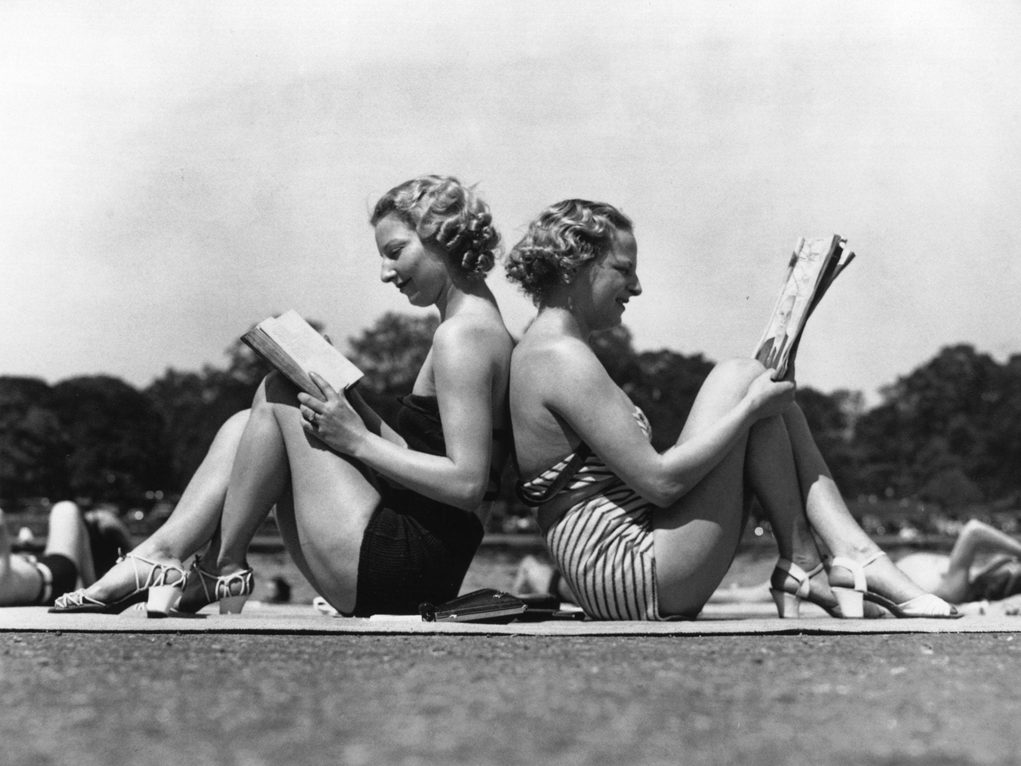 Two London women reading back to back as they sunbathe by the banks of the Serpentine in Hyde Park, London.