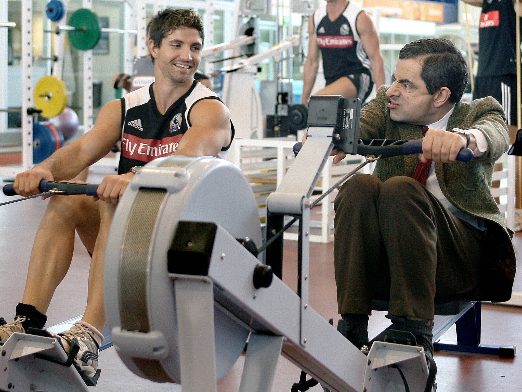 Actor Rowan Atkinson in character as Mr Bean trains on a rowing machine as Brodie Holland of the Magpies looks on during the Collingwood Magpies AFL gym training session at the Lexus Centre on March 9, 2007 in Melbourne, Australia.