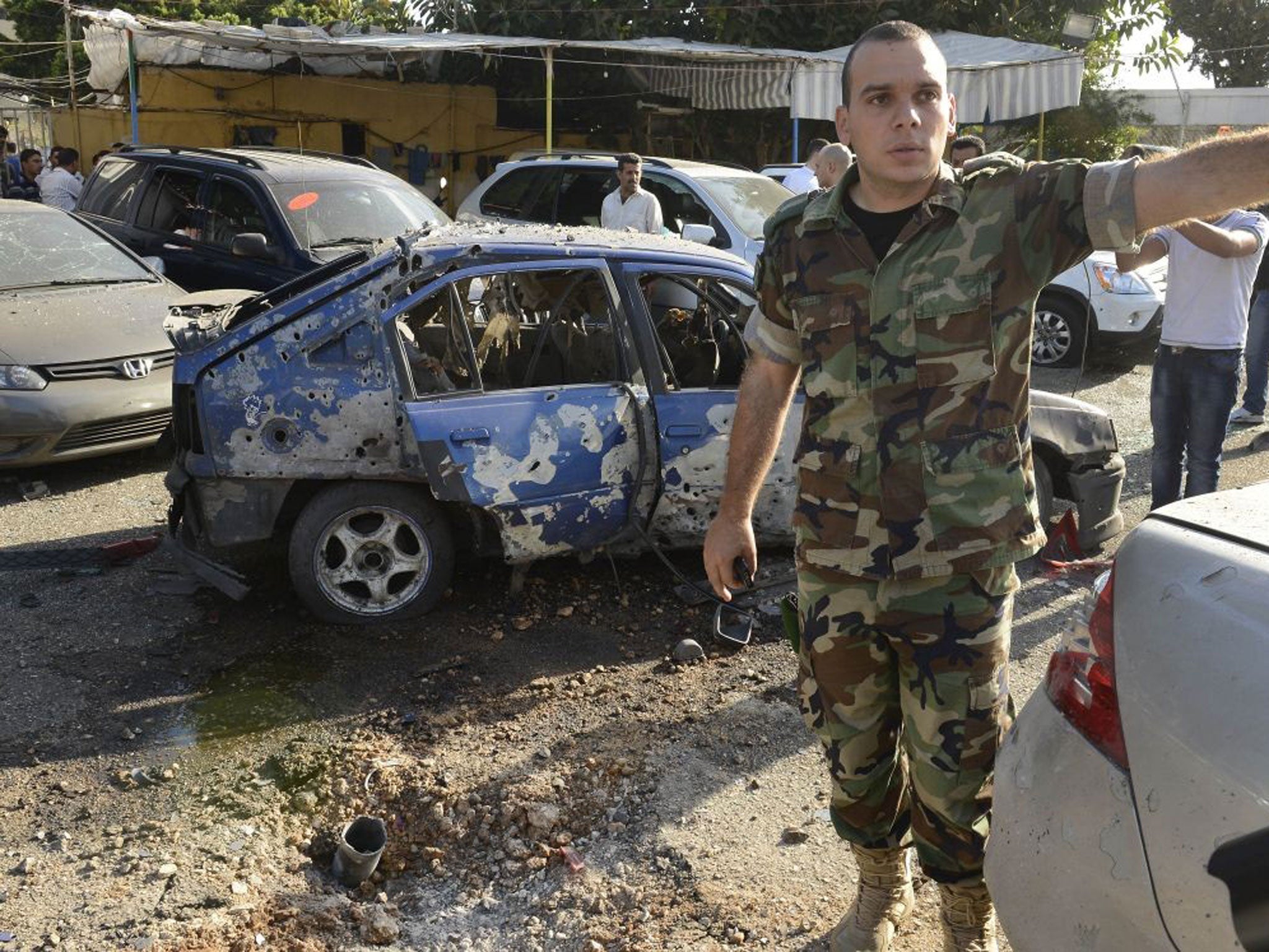 A Lebanese army officer stands next to a damaged car as he asks journalists to step back, at the scene where a rocket struck a car exhibit