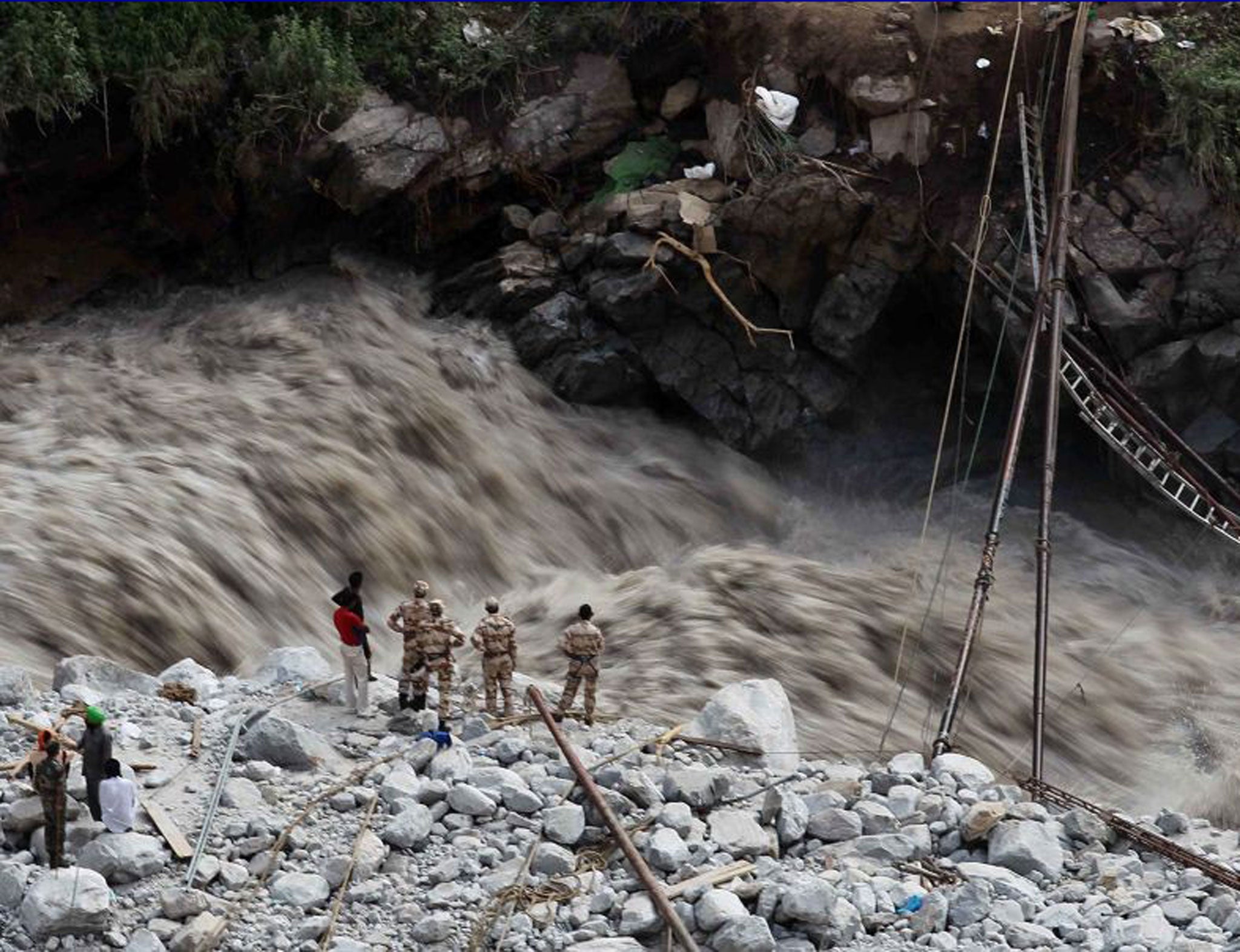 Water gushes down a river as Indian paramilitary soldiers and volunteers stand near a makeshift bridge