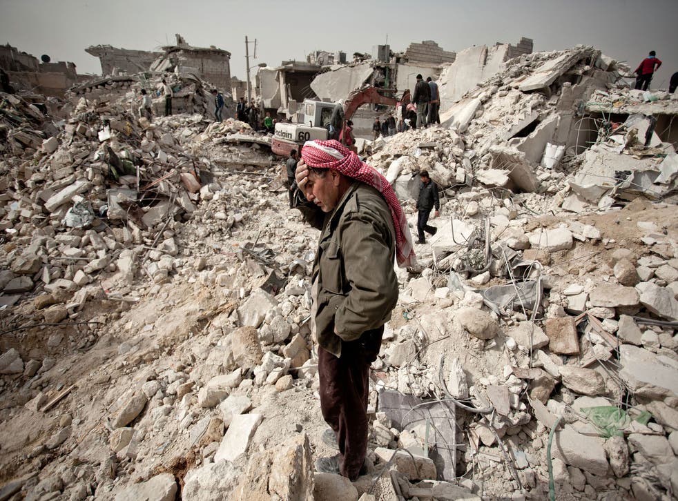 A Syrian man reacts while standing on the rubble of his house while others look for survivors