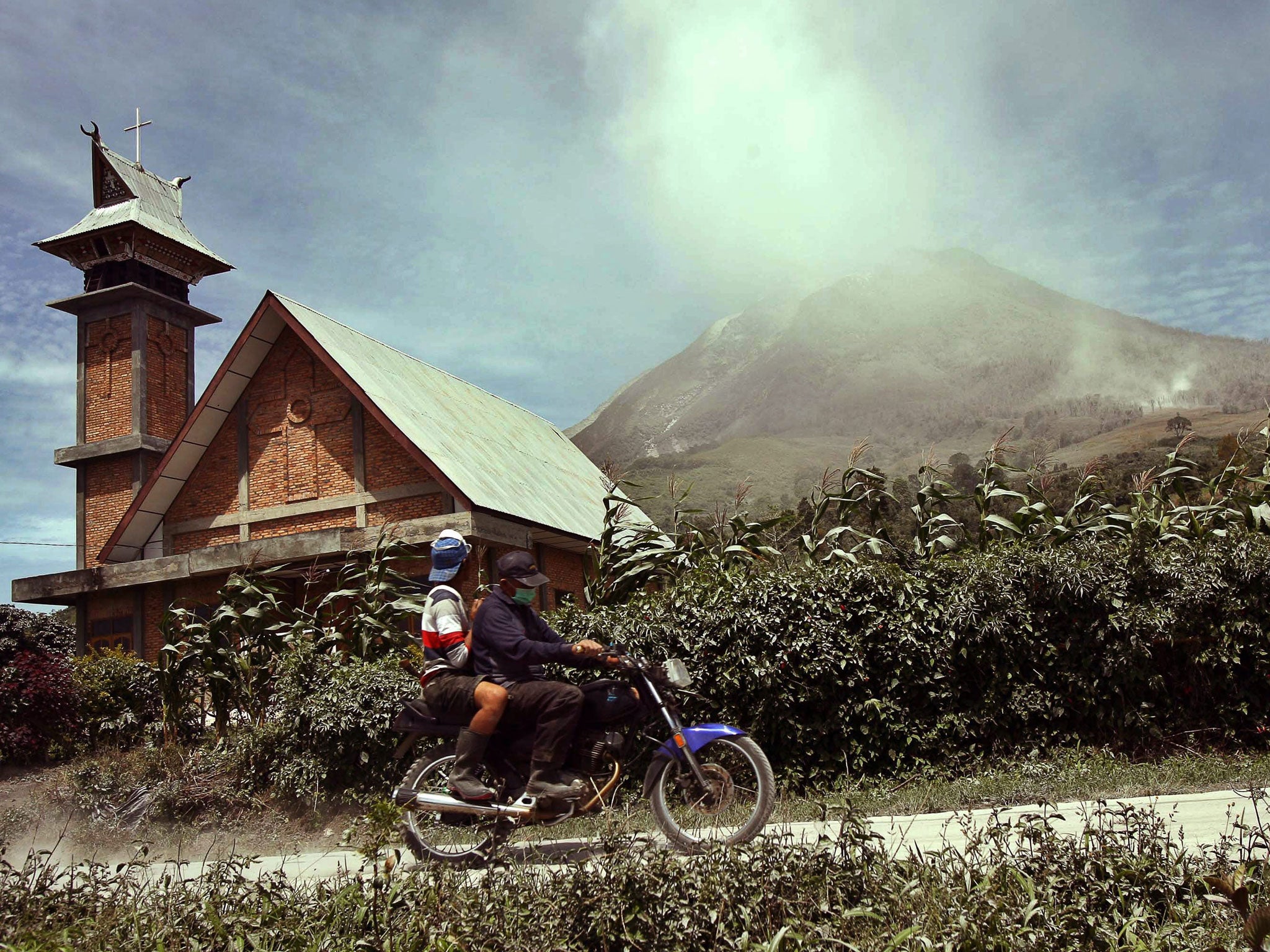 Indonesian villagers ride a motorbike as Mount Sinabung spews hot gas and ashes in the background during an eruption in Karo, North Sumatra province, Indonesia