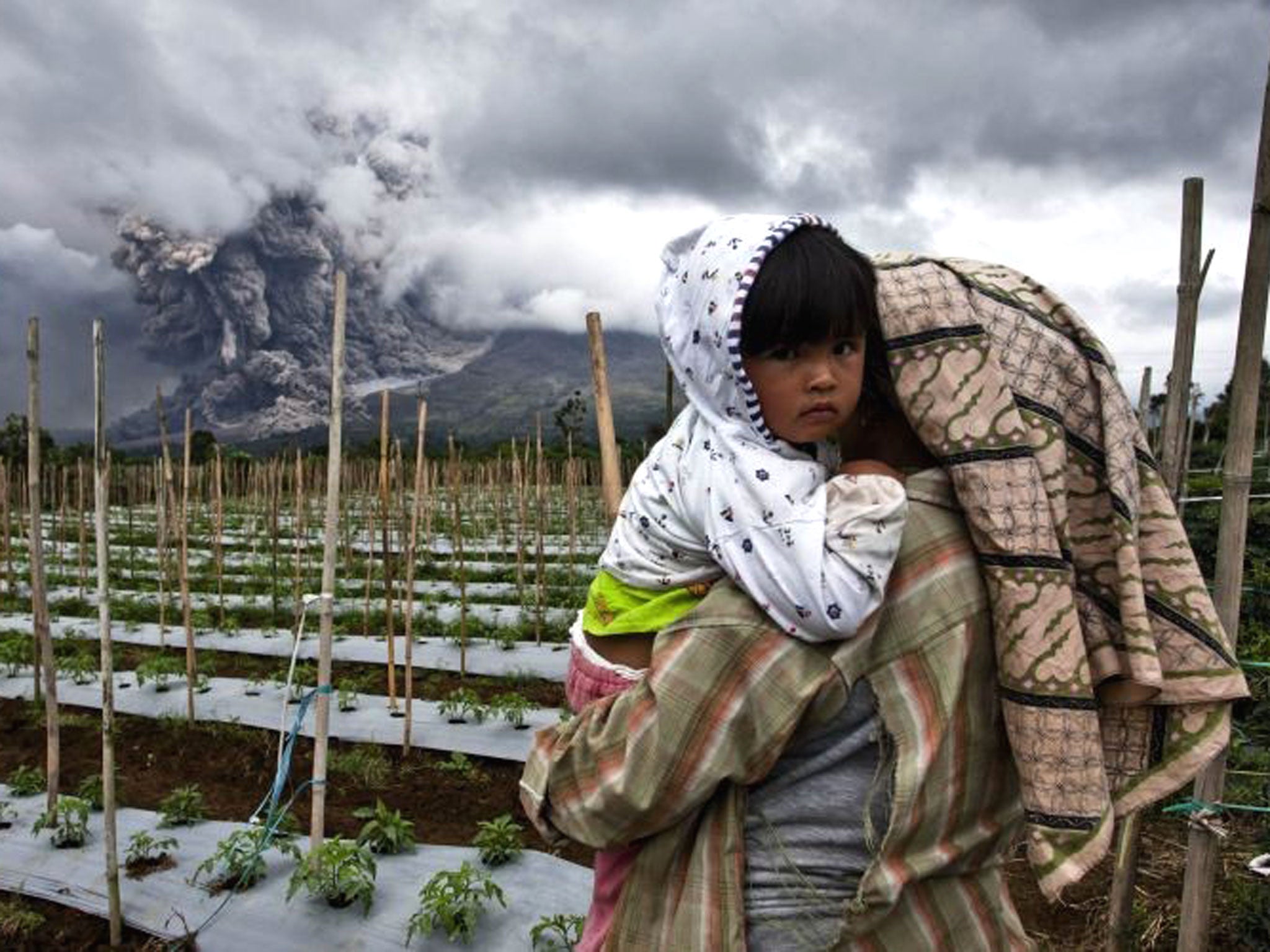A woman carries her daughter in a nearby field as Mount Sinabung spews pyroclastic smoke in Karo District, North Sumatra
