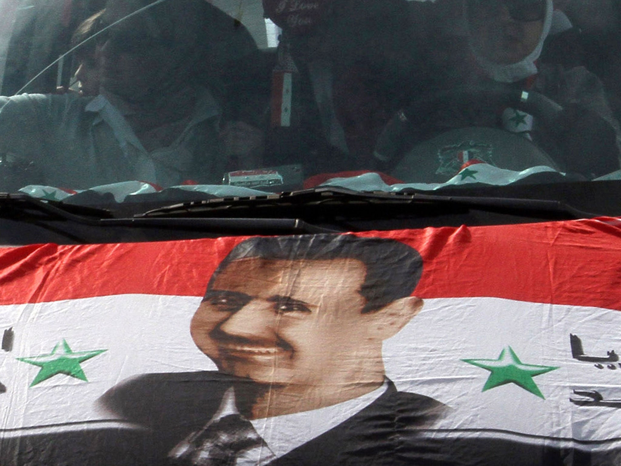 Syrians drive a car with a national flag on the front hood bearing a portrait of Syrian President Bashar al-Assad