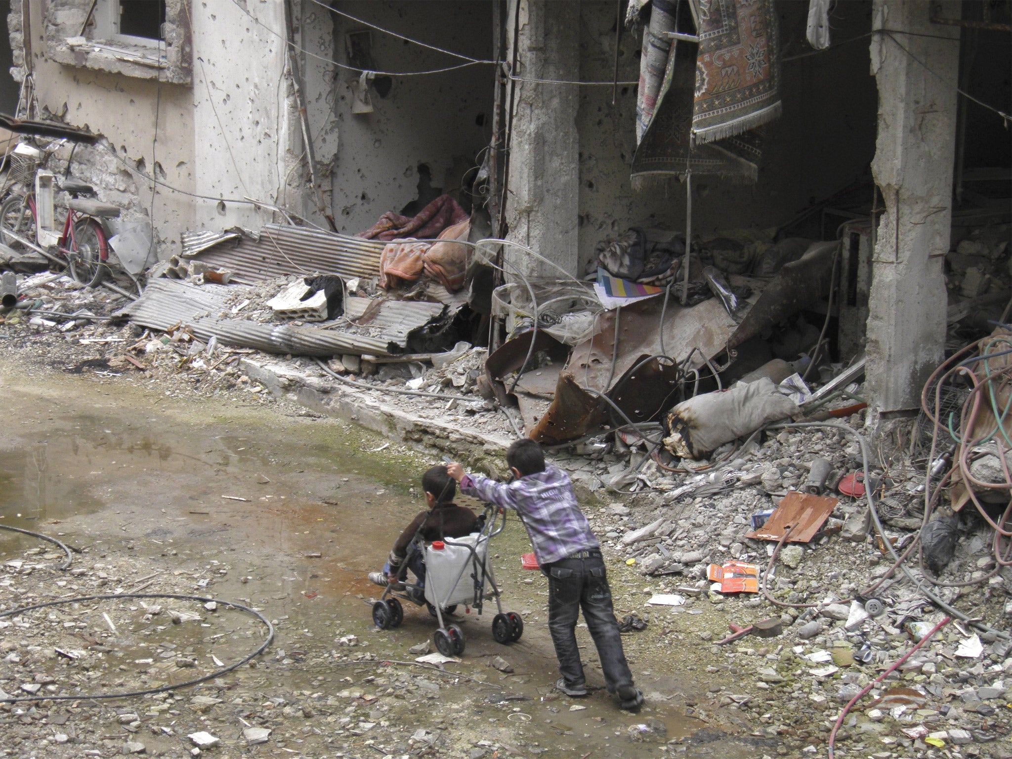 A boy pushes another on a pram along a damaged street in Homs