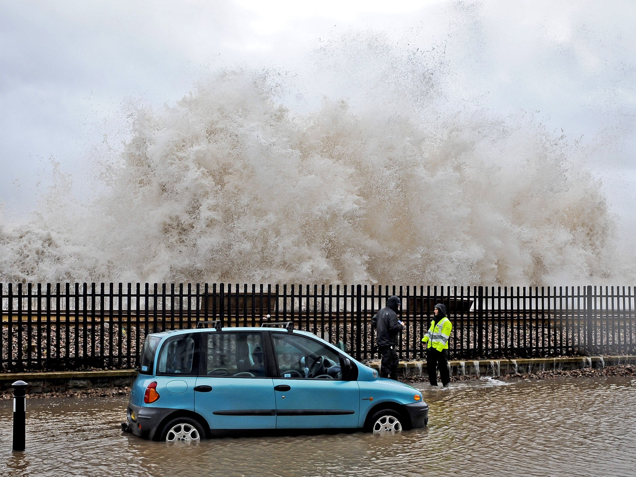 A huge wave breaks behind a car, which sits in seawater on a flooded street by the sea wall railway in Dawlish