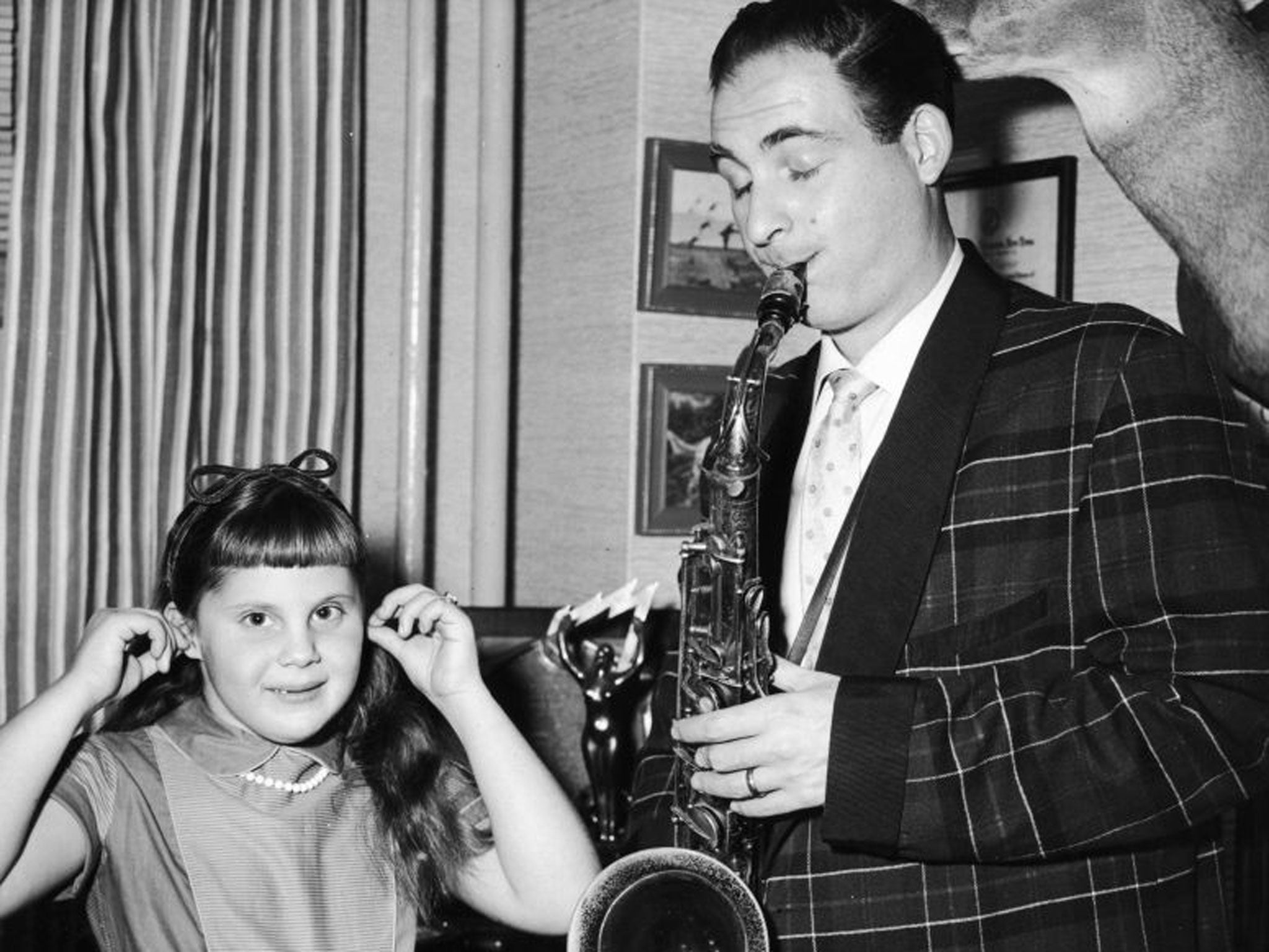 American comedian and actor Sid Caesar plays the saxophone indoors as his daughter, Karen, holds her ears.