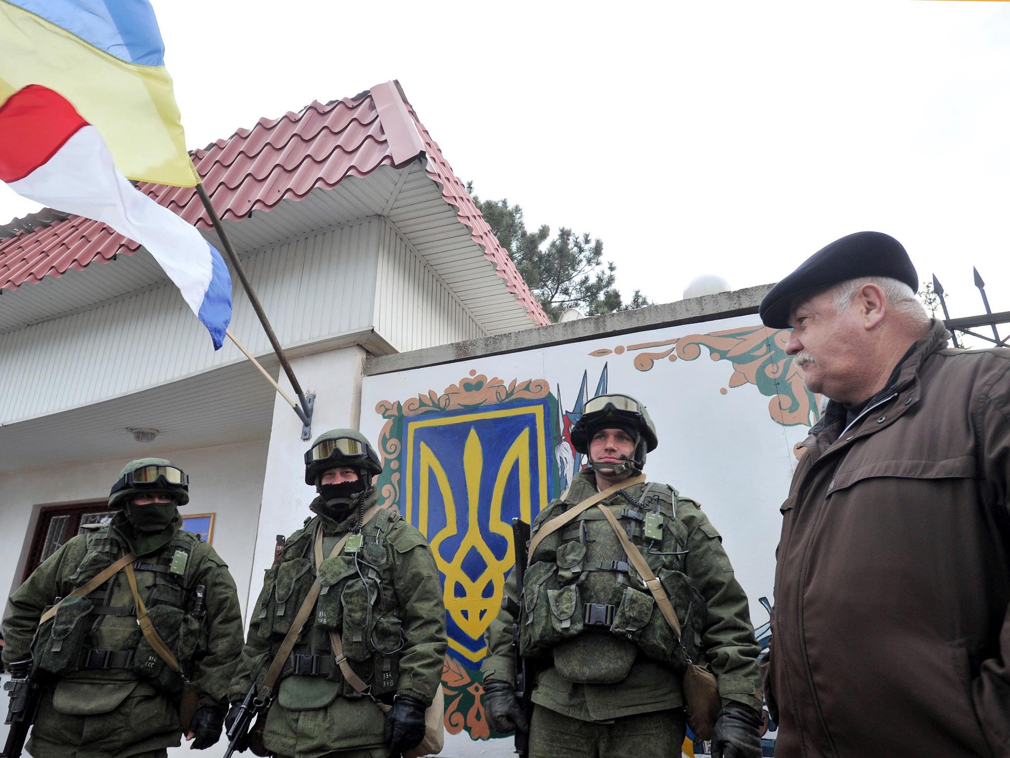 An Autonomous Republic of Crimea and a Ukrainian flag float side by side as a man watches armed men in military fatigues blocking access to a Ukrainian border guards base not far from the village of Perevalne near Simferopol