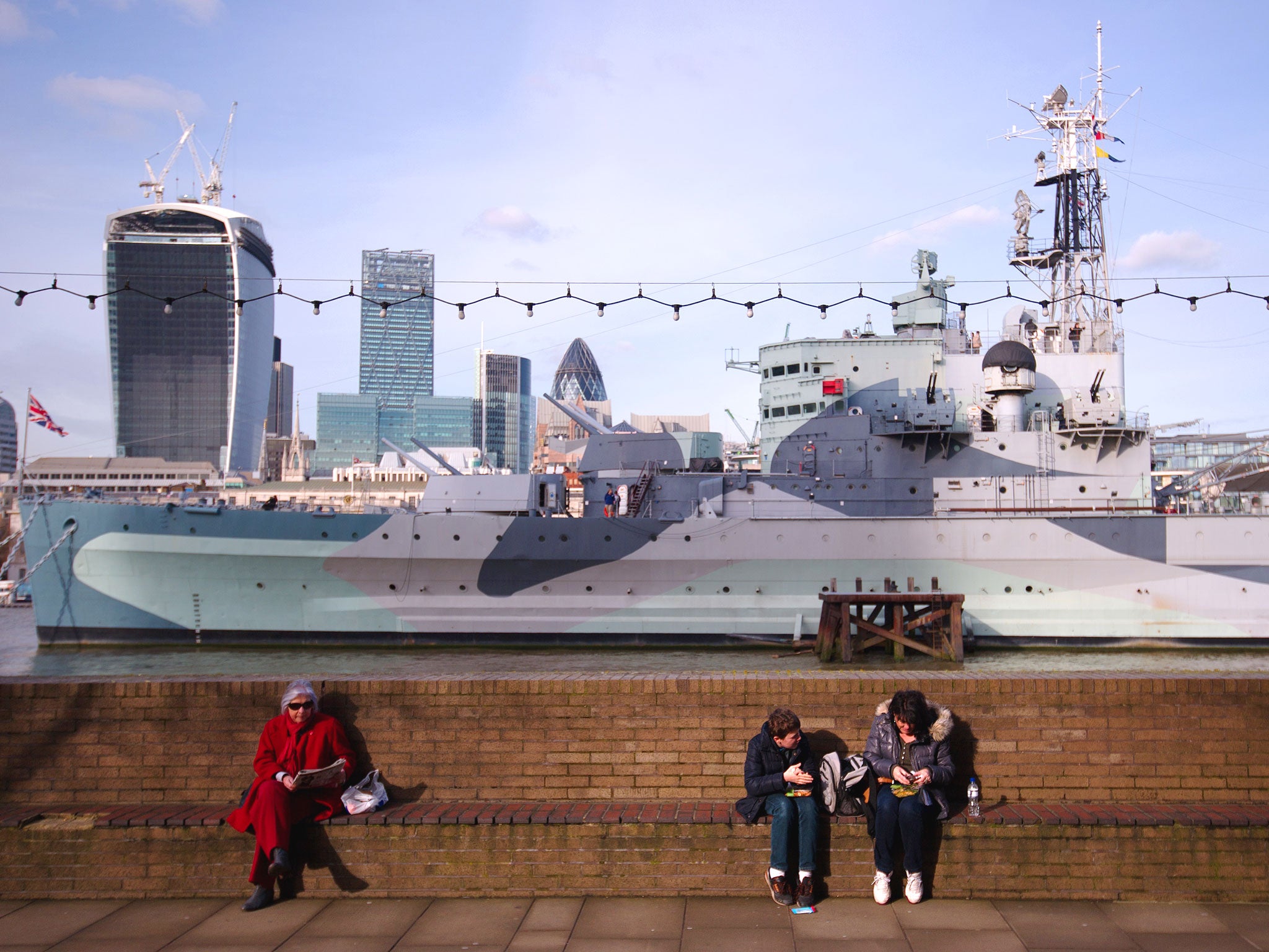 People sit in the sunshine with HMS Belfast on the River Thames in the background in central London