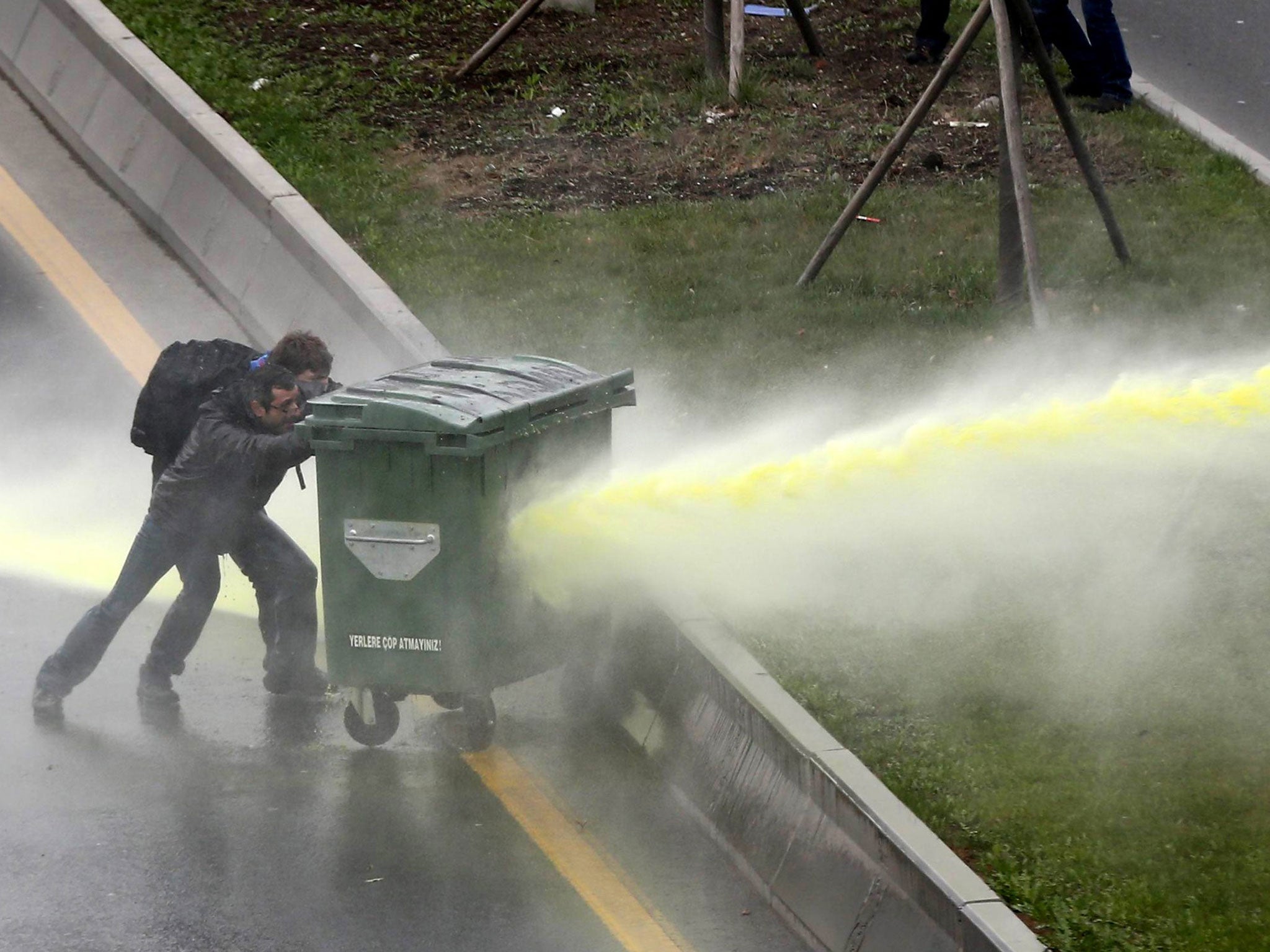People protect themselves behind a wheelie bin as anti-riot police fires water cannon in Turkey