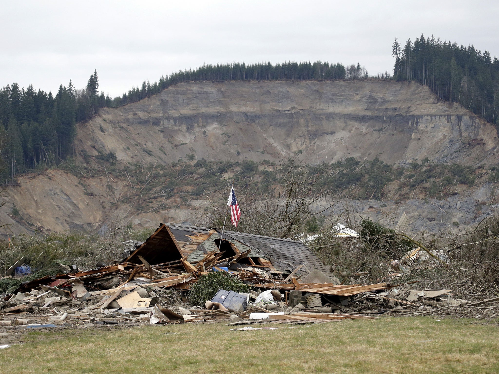 A flag, put up by volunteers helping search the area, stands in the ruins of a home left at the end of a deadly mudslide from the now-barren hillside seen about a mile behind in Oso