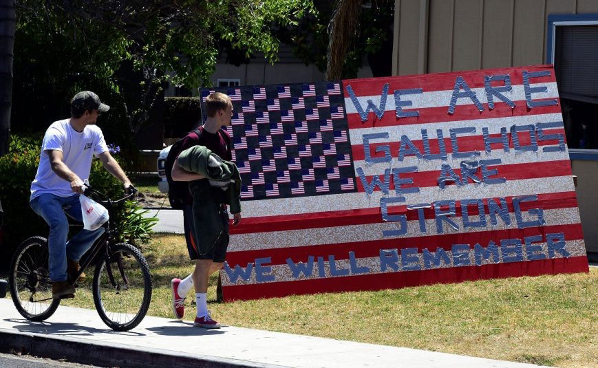 Two students look at a sign that reads "We are Gauchos, We are Strong, We will Remember" set up outside an apartment in the college town of Isla Vista, California