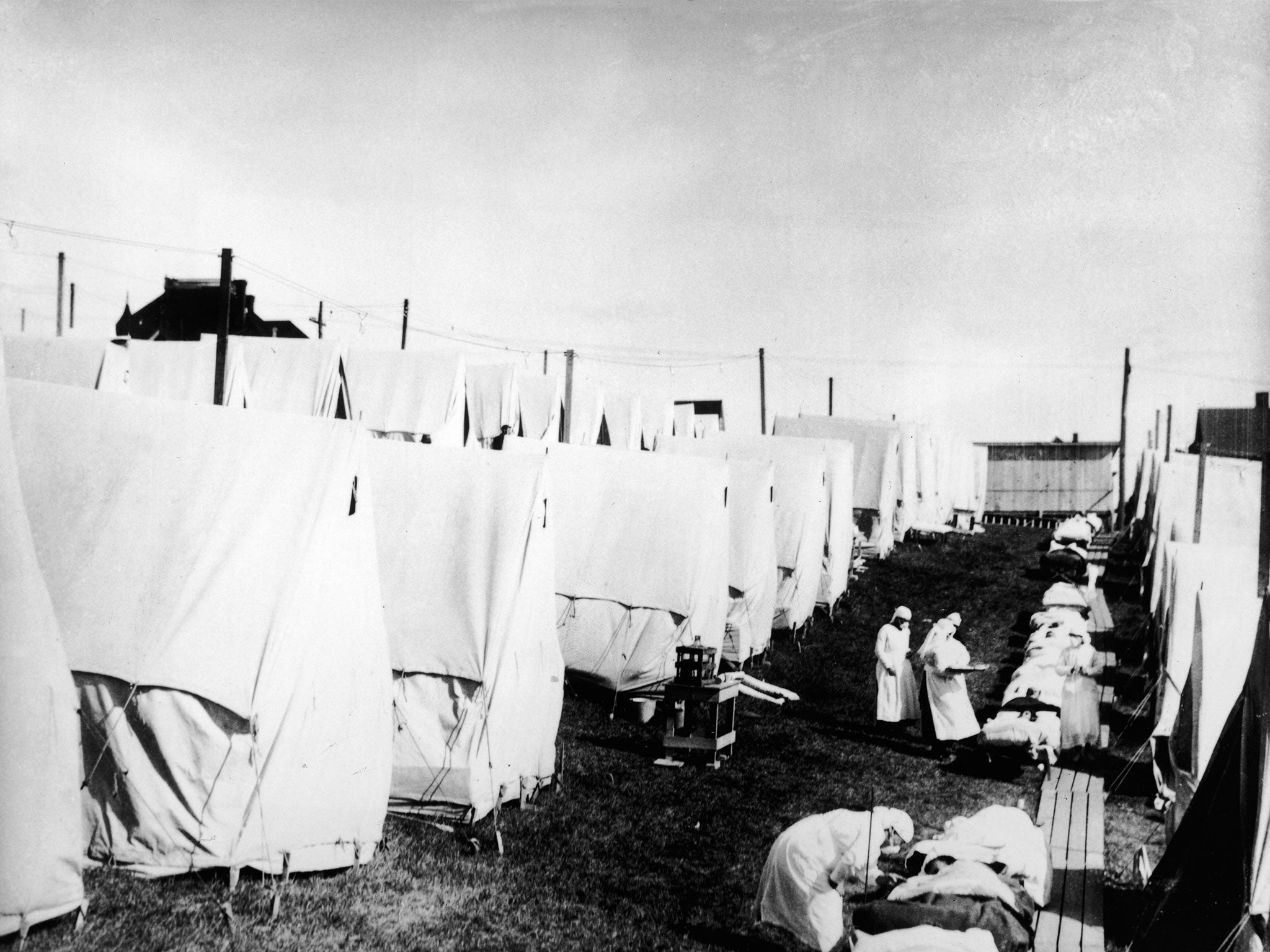 Masked doctors and nurses treat flu patients lying on cots and in outdoor tents at a hospital camp during the influenza epidemic of 1918