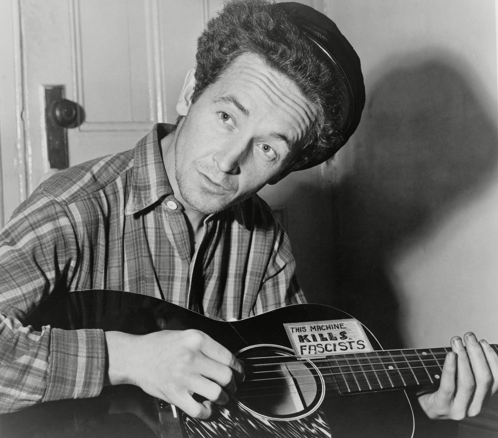 Woody Guthrie with his guitar. The sticker reads 'This machine kills fascists'