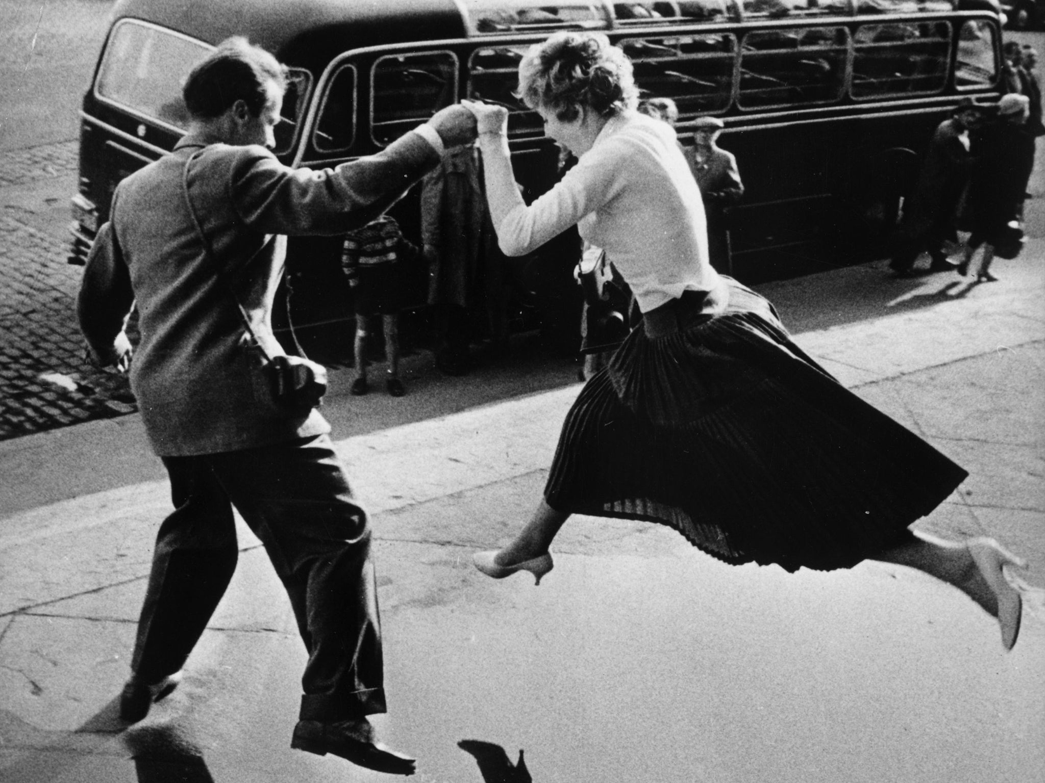 A man gives a woman a helping hand as she takes a flying leap over a large puddle on the pavement