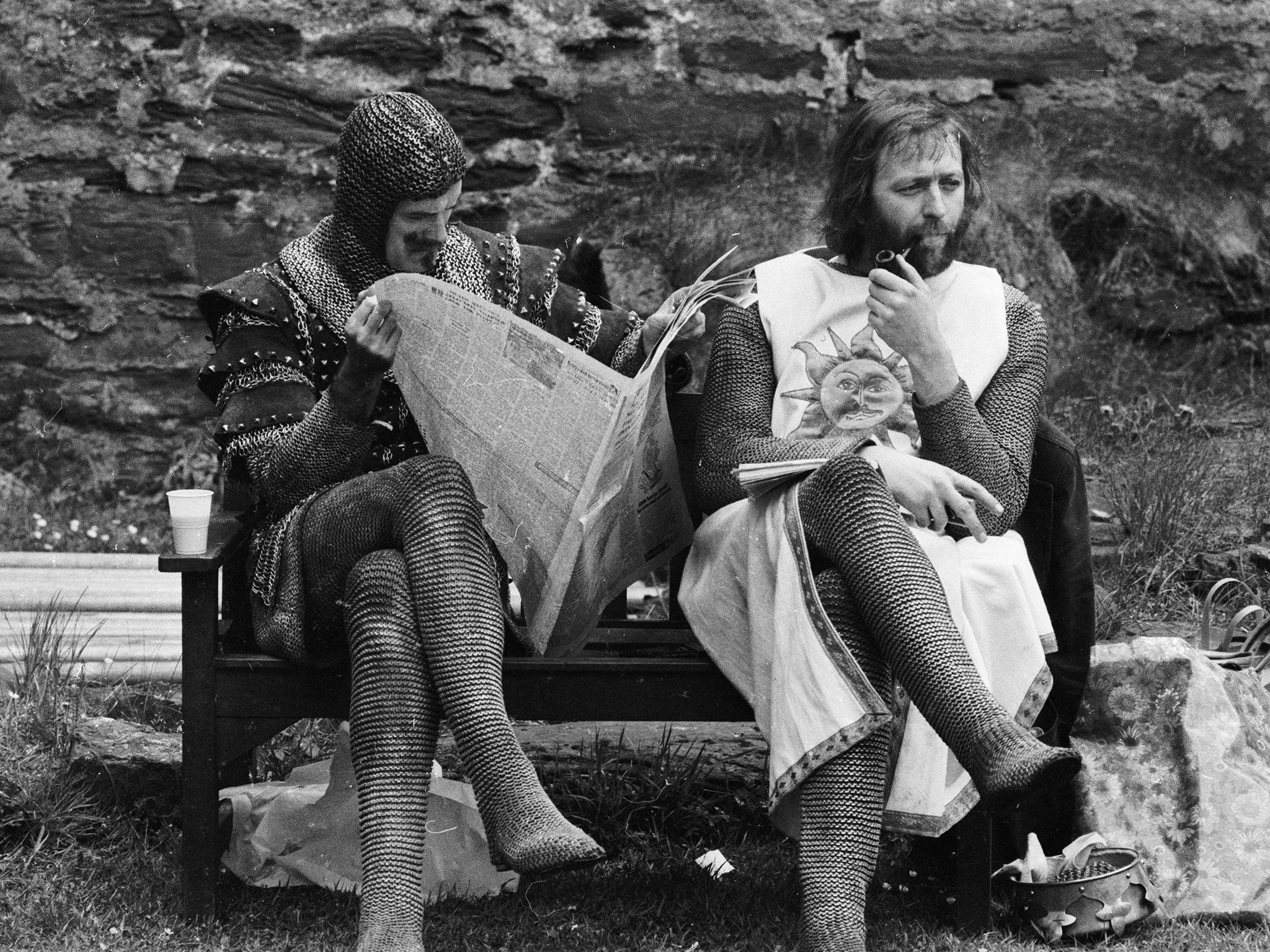 A chainmail-clad John Cleese reads a newspaper while Graham Chapman smokes a quiet pipe on the set of Monty Python and the Holy Grail in 1974