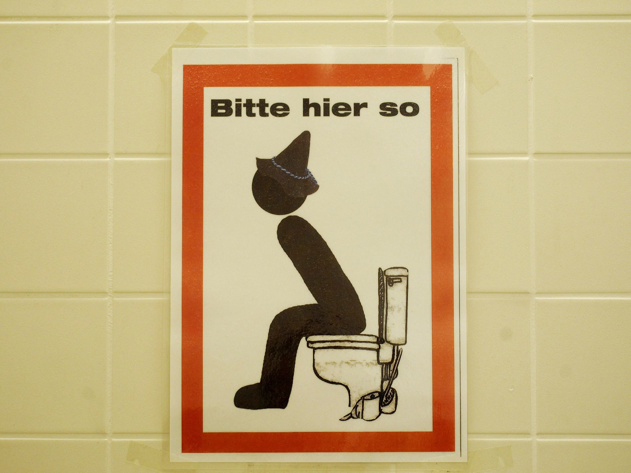 A sign reading "Bitte hier so" (please like this here) and depicting a seated man with bavarian hat on a toilet