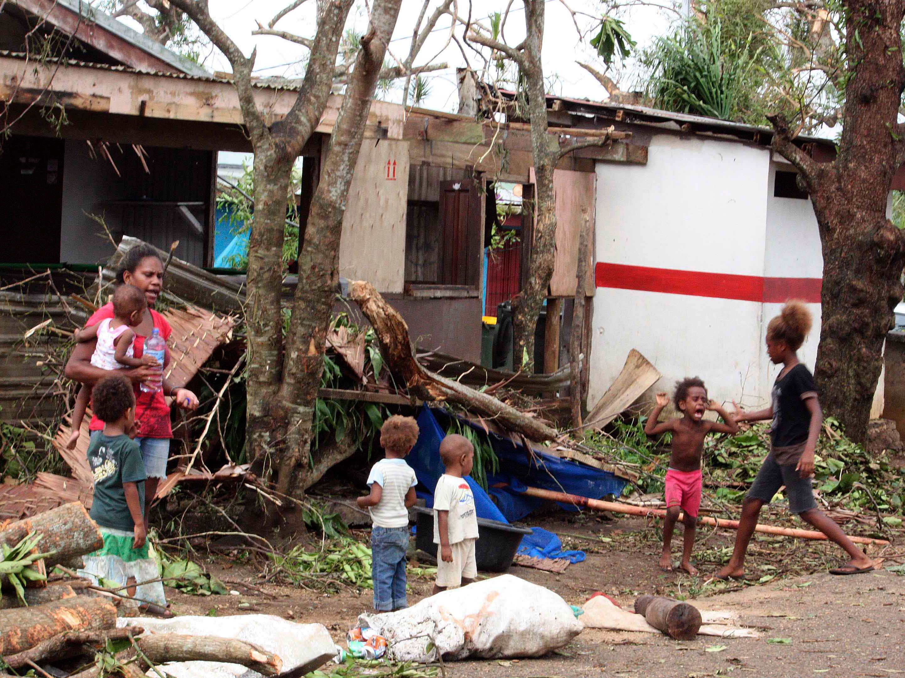 A woman carrying a baby stands with children outside homes damaged by Cyclone Pam, on a street surrounded by debris in Port Vila