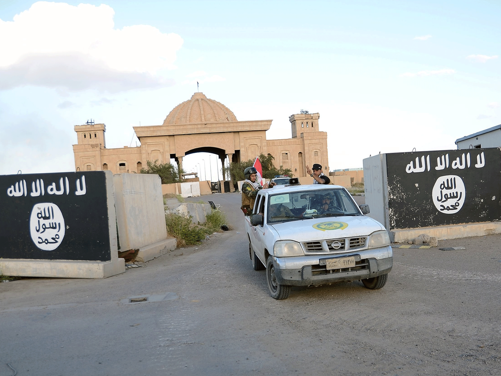 Shia paramilitary fighters ride past a wall painted with the black flag commonly used by Isis militants, with the palaces of former Iraqi president Saddam Hussein behind them, in Tikrit, March 31, 2015
