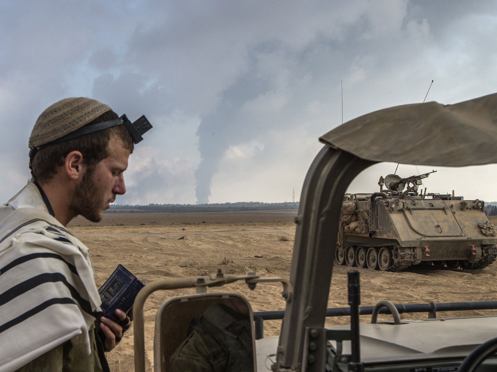 An Israeli soldier prays on the Israeli side of the border with the Gaza Strip, on July 29, 2014, as smoke billows from a power plant following overnight Israeli shelling in the coastal Palestinian enclave.