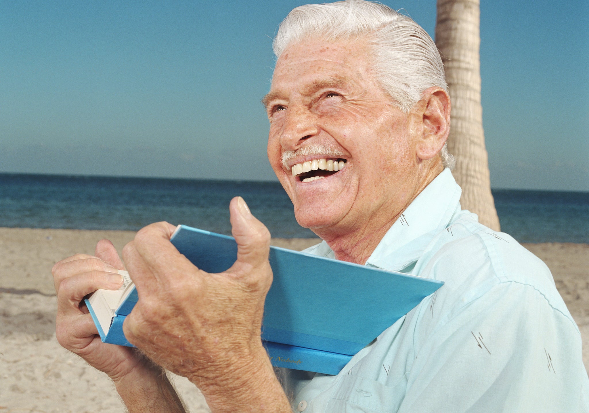 A senior white man reading on the beach, laughing.