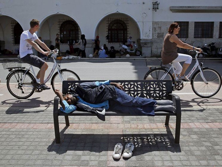 An Afghan immigrant sleeps on a bench outside a local police station as tourists cycle by