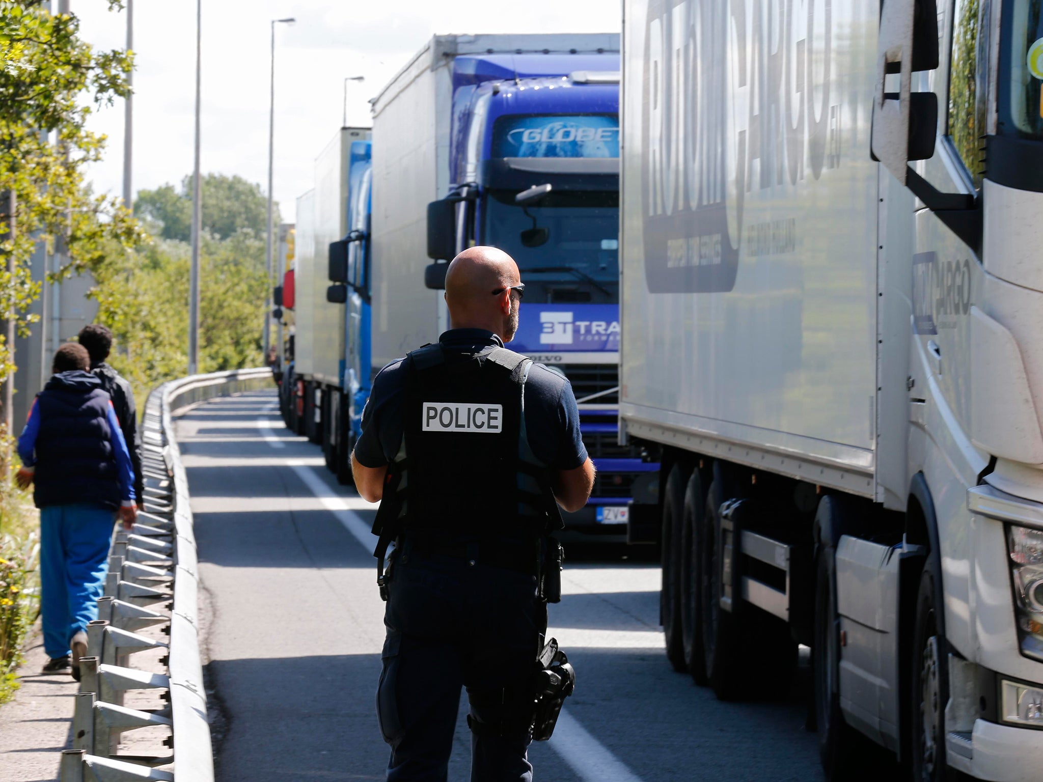 Migrants walk along the roadside while a French policeman secures the area as lorries queue in Calais