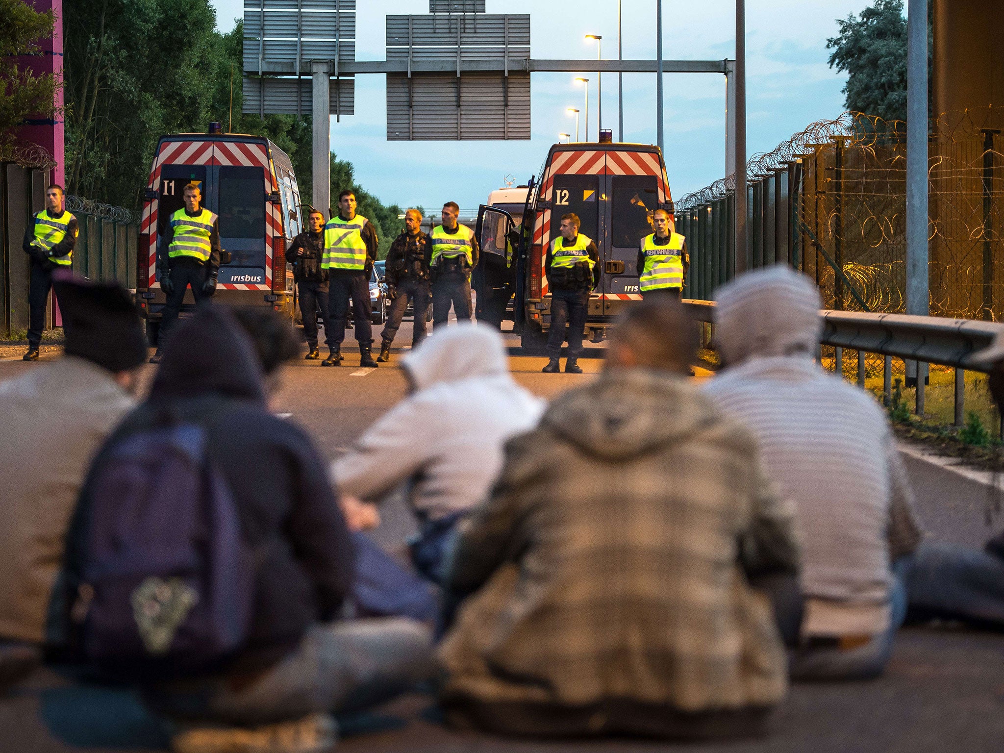 French gendarmes stand facing a group of seated migrants as they block their way in the Eurotunnel site
