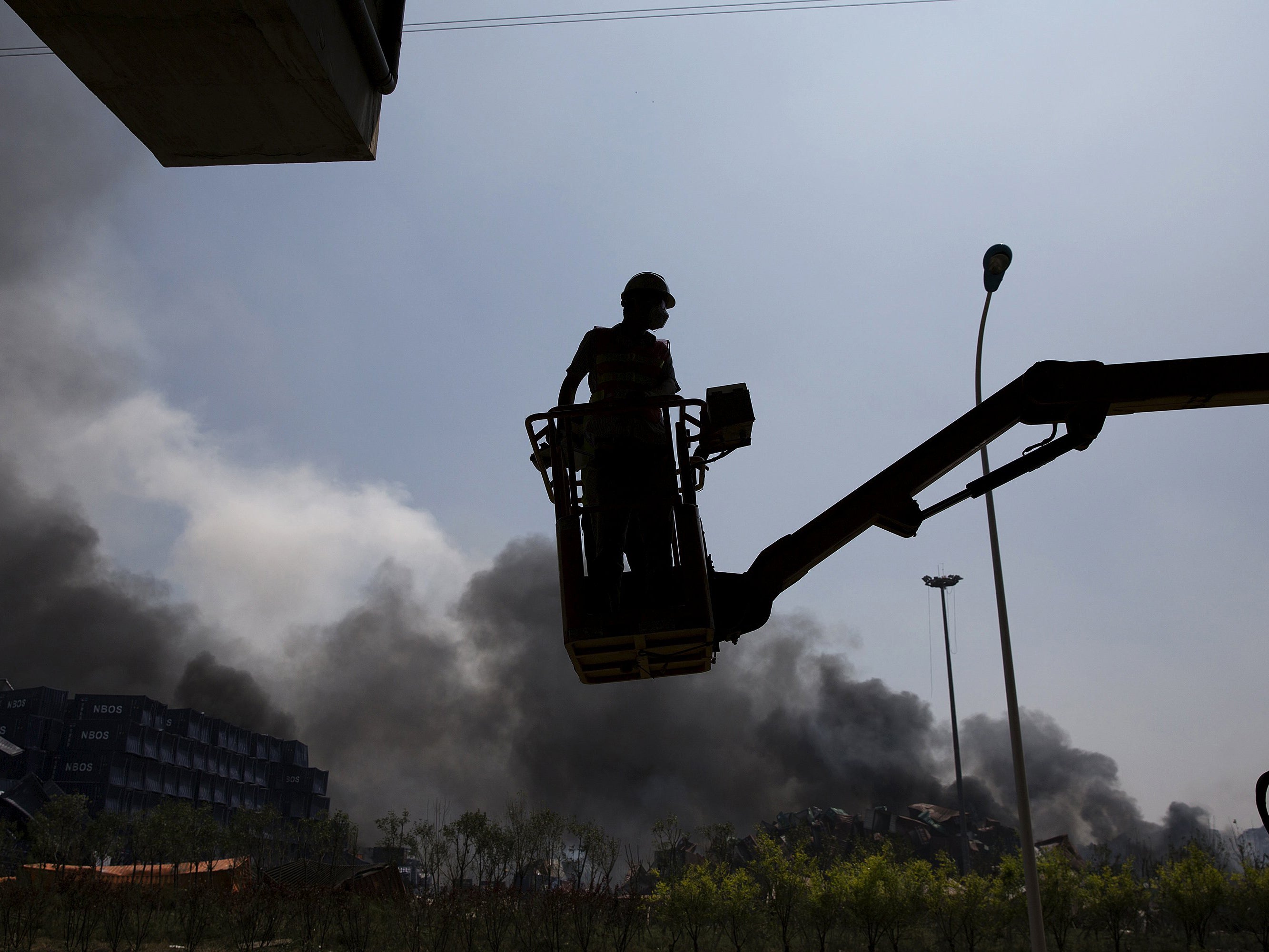 A emergency worker is lifted by a crane as smokes plumes from the explosion site in Binhai new district in Tianjin