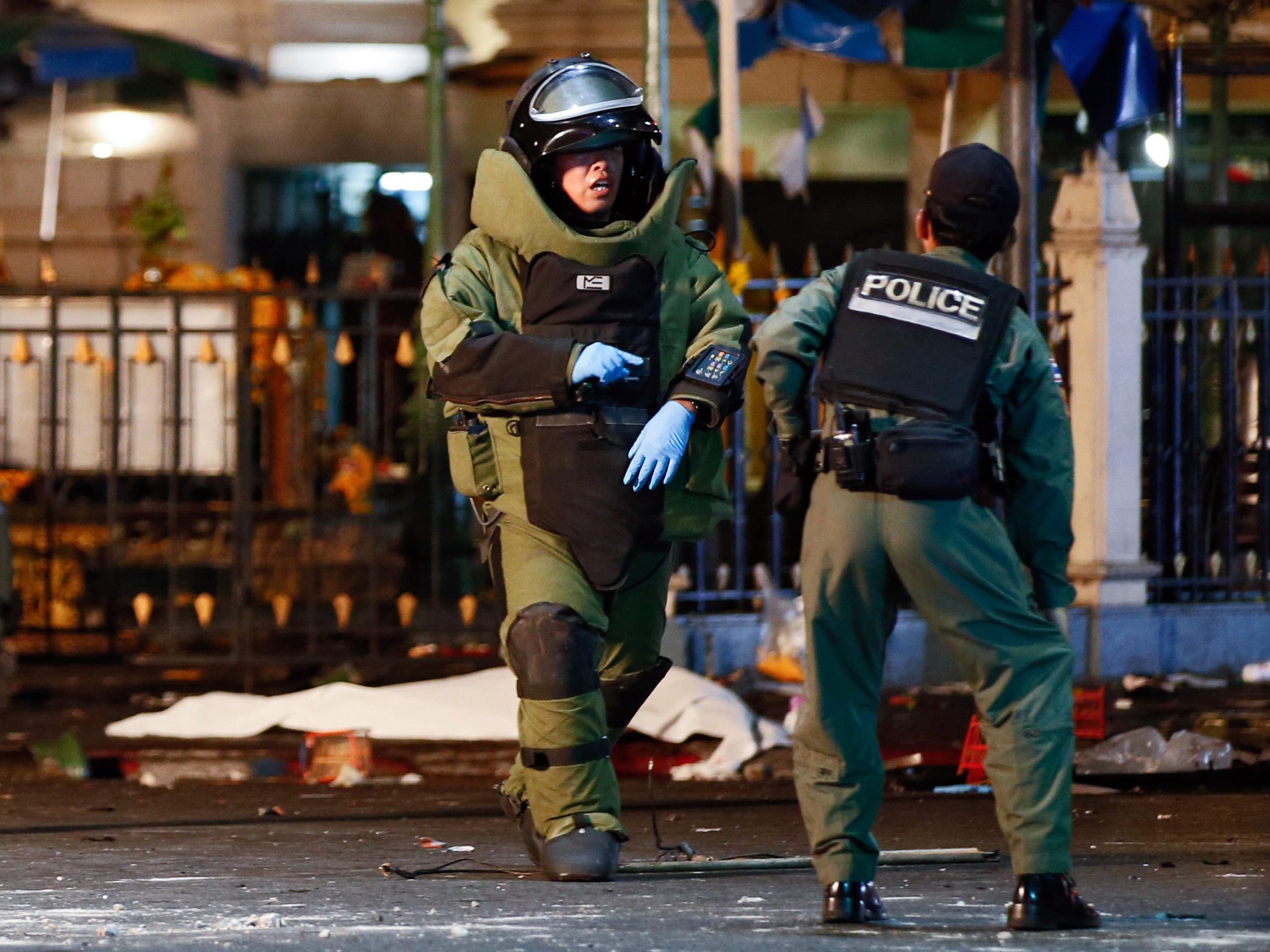 A member of the bomb squad in protective suit talks to a colleague as the body of a victim is seen on the background