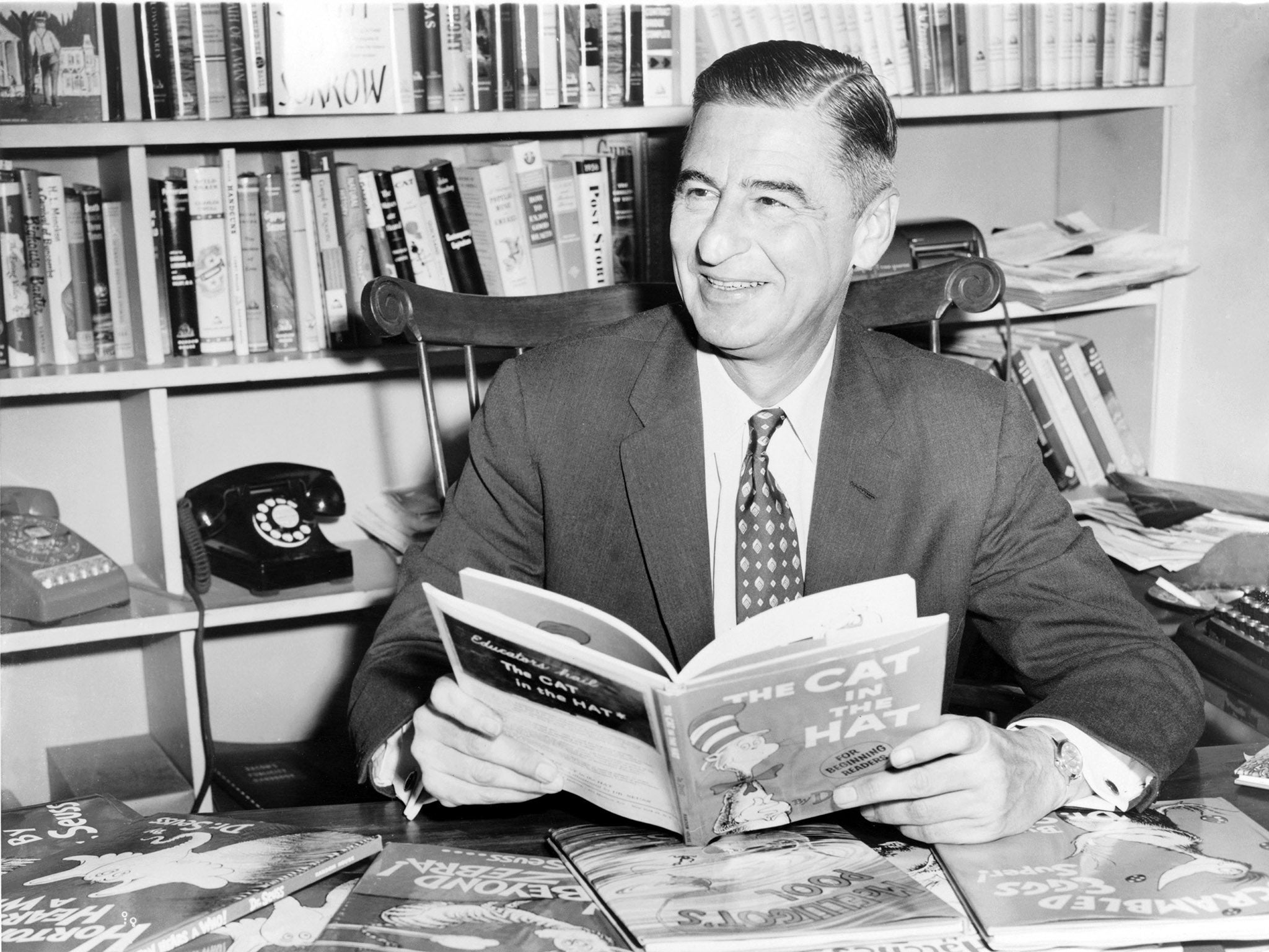 Ted Geisel (Dr Seuss) seated at desk covered with his uniquely humorous children's books