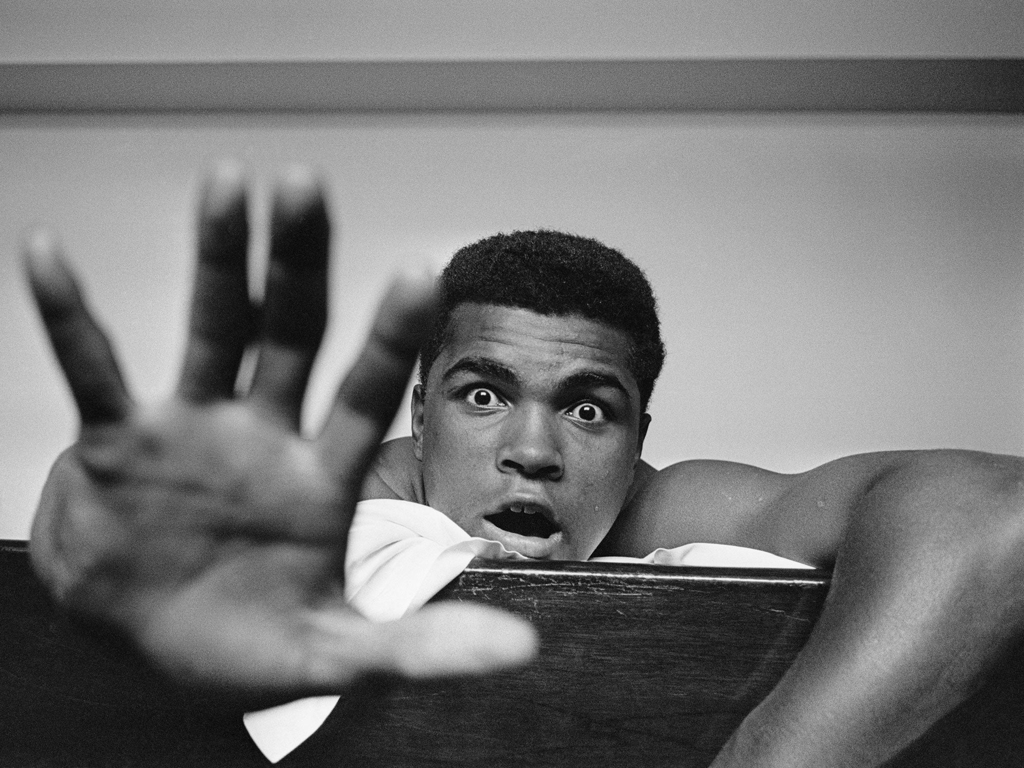 Muhammad Ali lying on his hotel bed in London, 27 May, 1963. He holds up five fingers in a prediction of how many rounds it will take him to knock out British boxer Henry Cooper