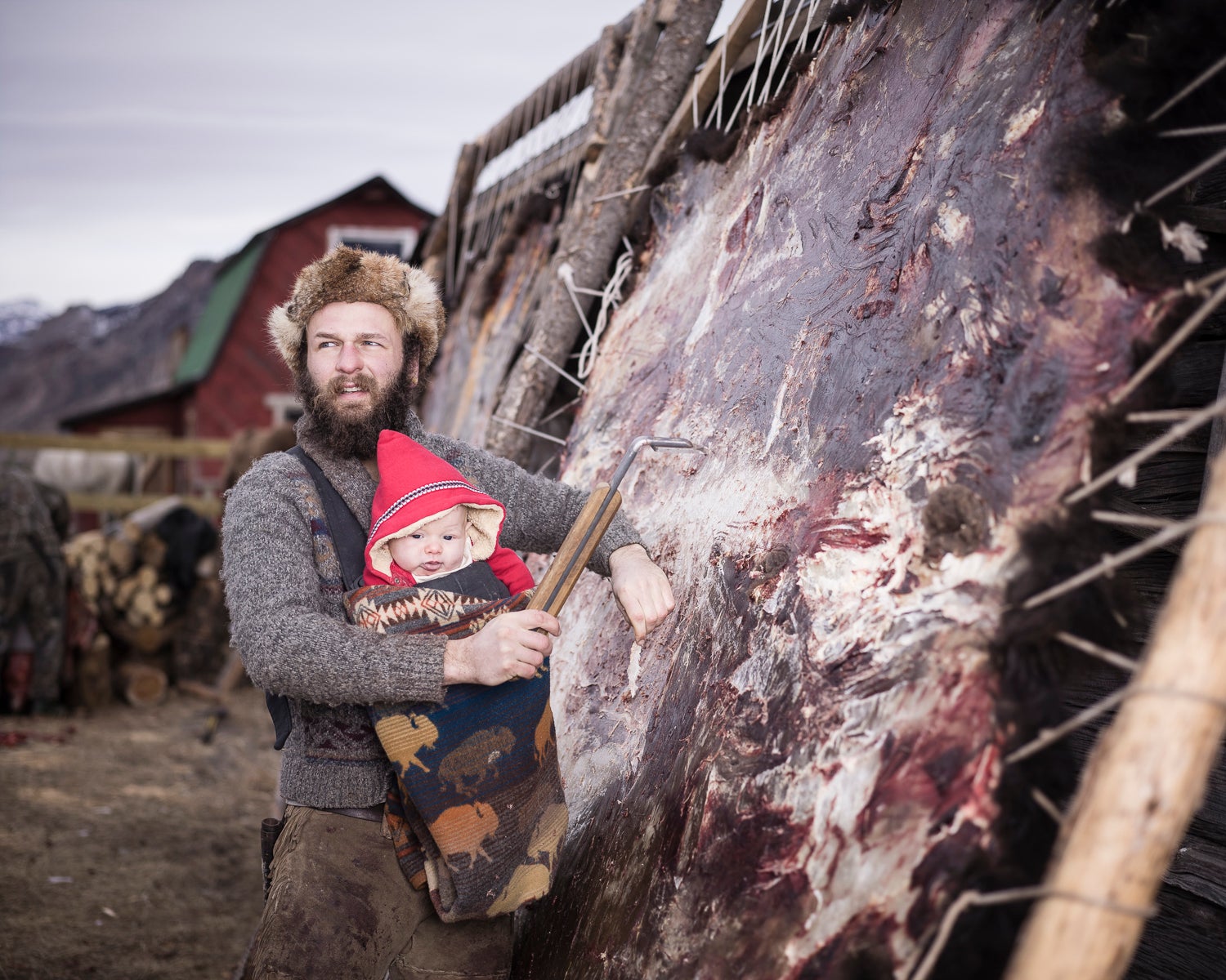 Alex carries his son as he works at fleshing a hide for tanning