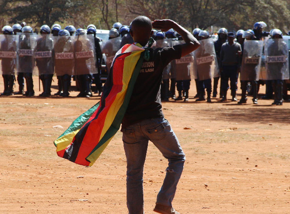 Riot police watch a man with a Zimbabwean flag over his shoulders saluting during a protest