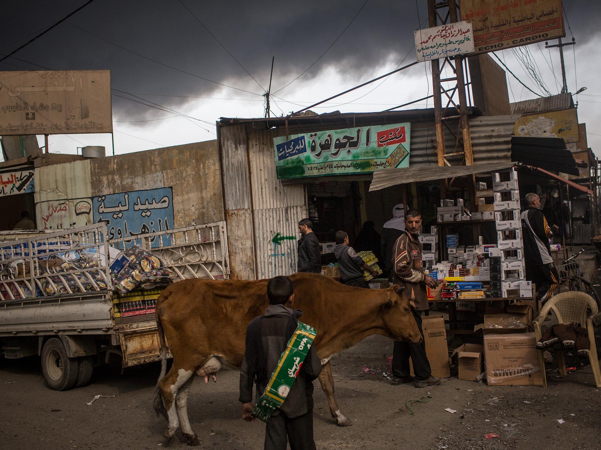 Black plumes of smoke hang over the street market; locals stand idle as a cow wanders by