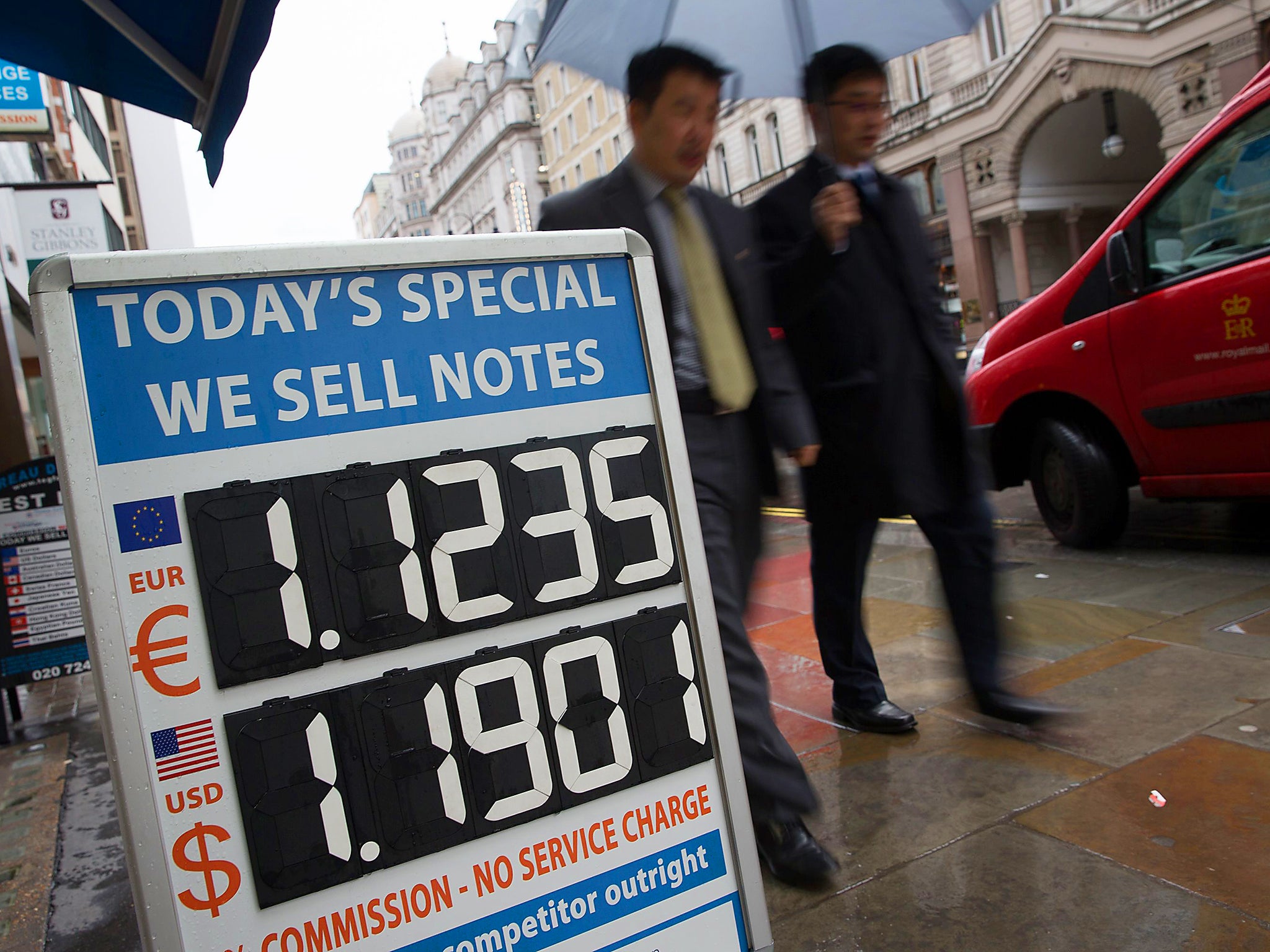 Pedestrians walk past a board displaying the price of Euro and US dollars against British pound Sterling