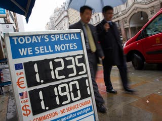 Pedestrians walk past a board displaying the price of Euro and US dollars against British pound Sterling