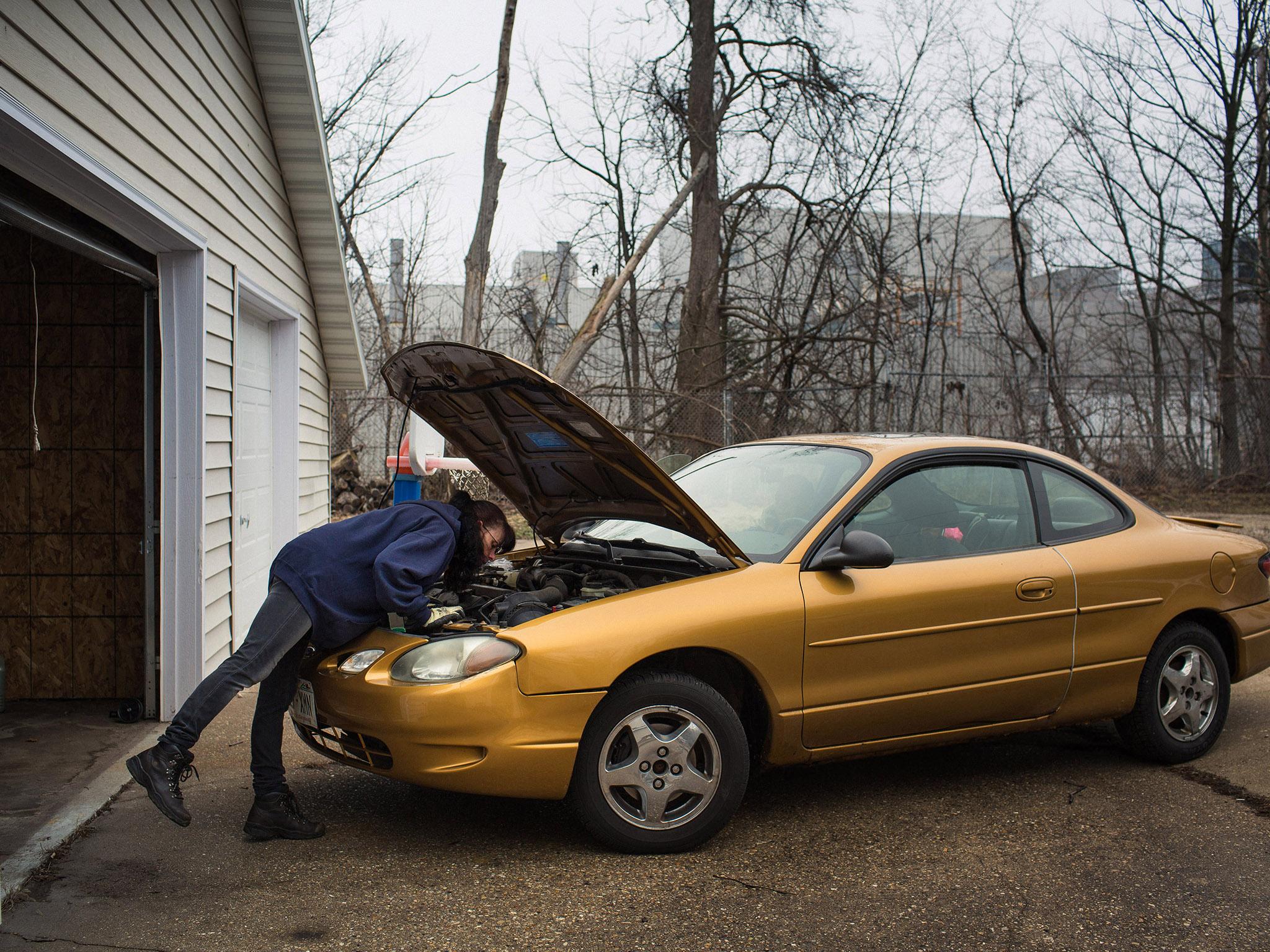 With the decommissioned assembly plant visible behind her, Mary Johnston tries to fix the oxygen sensor on her Ford Taurus. Her father taught her how to fix cars, she says, but she is missing the necessary wrench to finish the job