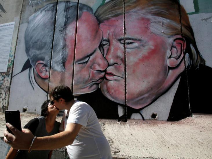 Tourists kiss each other as they stand in front of a mural depicting US President Donald Trump and Israel's Prime Minister Benjamin Netanyahu kissing each other in the West Bank city of Bethlehem