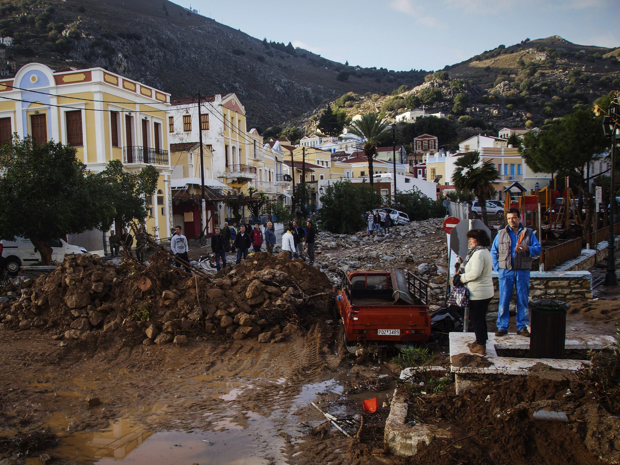 Residents stand next to a damaged road as a washed up vehicle is seen in the background following a powerful storm on the Greek island of Symi