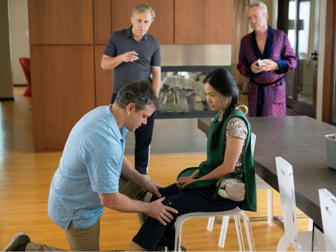 A scene from 'Downsizing' where Mat Damon is on one knee touching Hon Chau's knee with his hands as she is on a chair because she is an amputee. She is looking at Mat as he is facing at her legs.