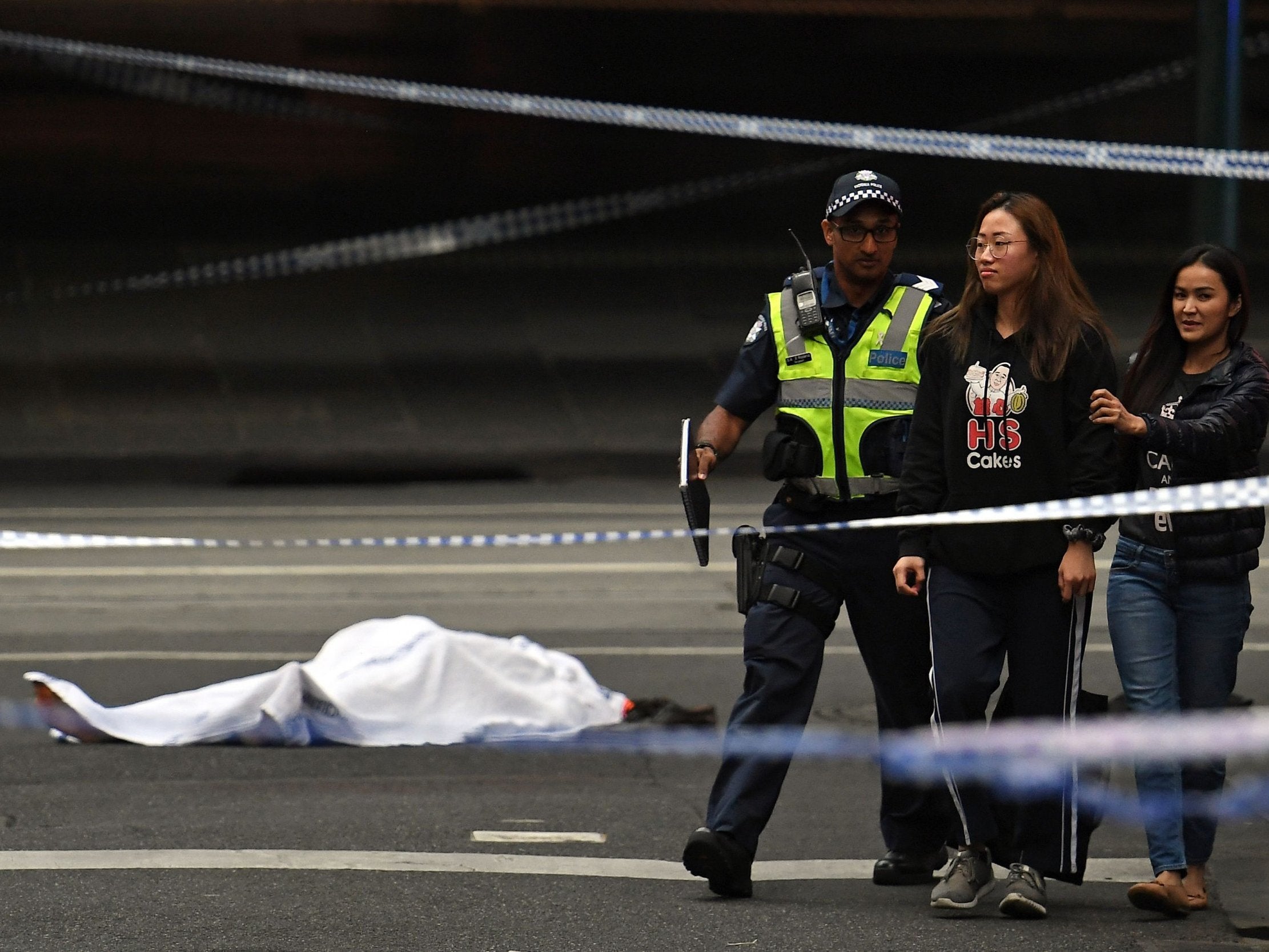 A police officer directs people away from the crime scene as a body is seen covered with a white sheet following a stabbing incident in Melbourne
