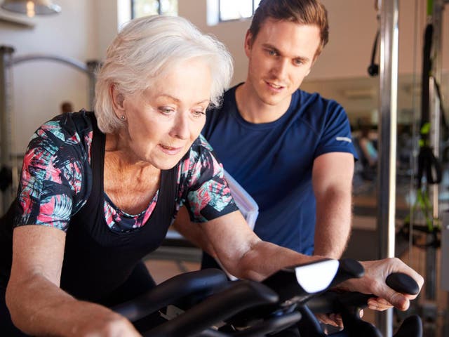 Senior woman exercising on cycling machine being encouraged by male personal trainer in gym
