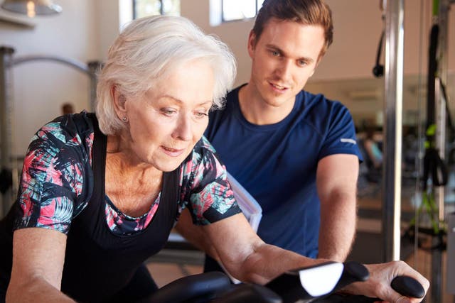 Senior woman exercising on cycling machine being encouraged by male personal trainer in gym